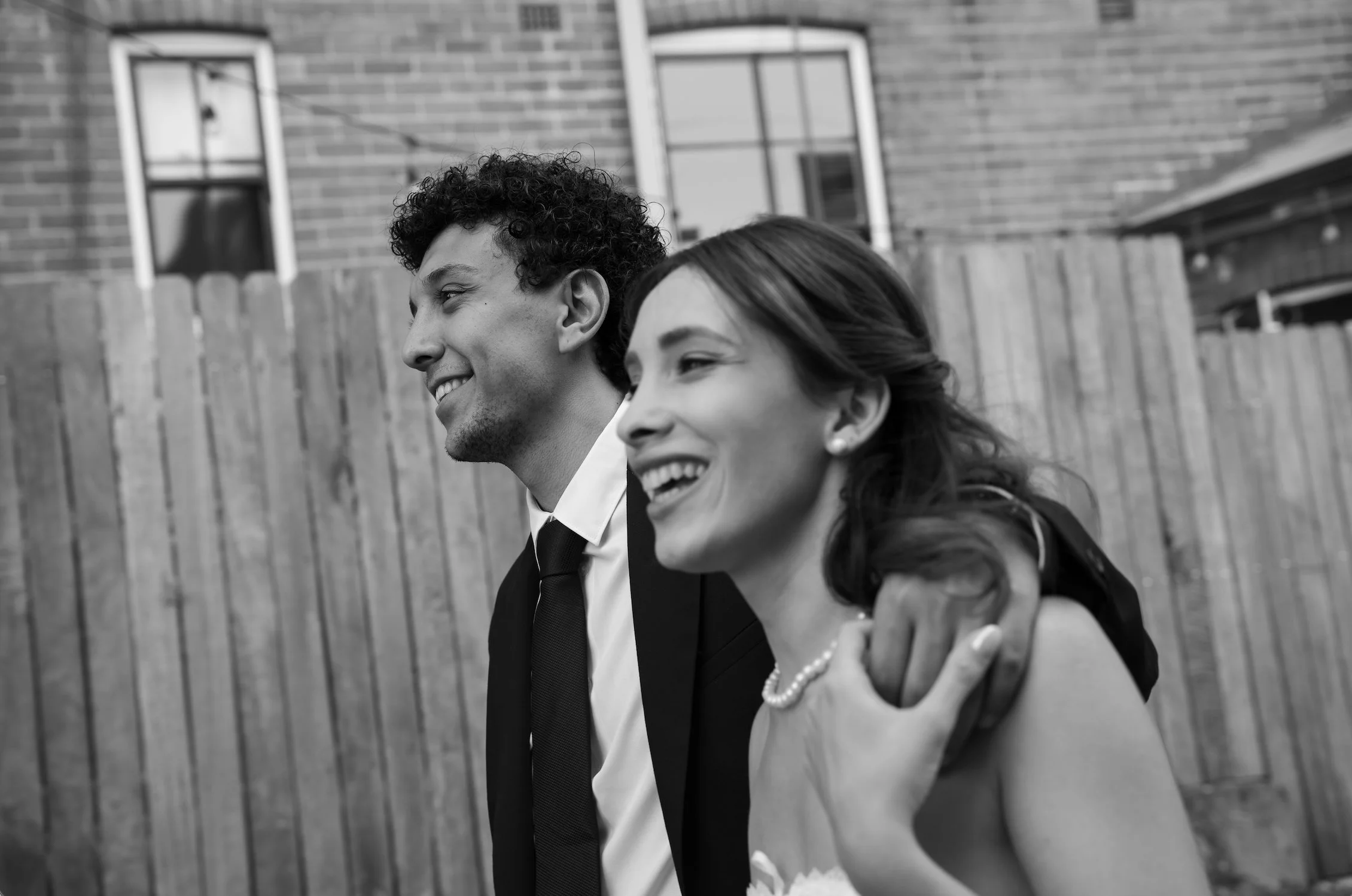 Black and white photo of a happy couple in formal attire, standing outdoors near a wooden fence and brick house, smiling and enjoying each other's company.