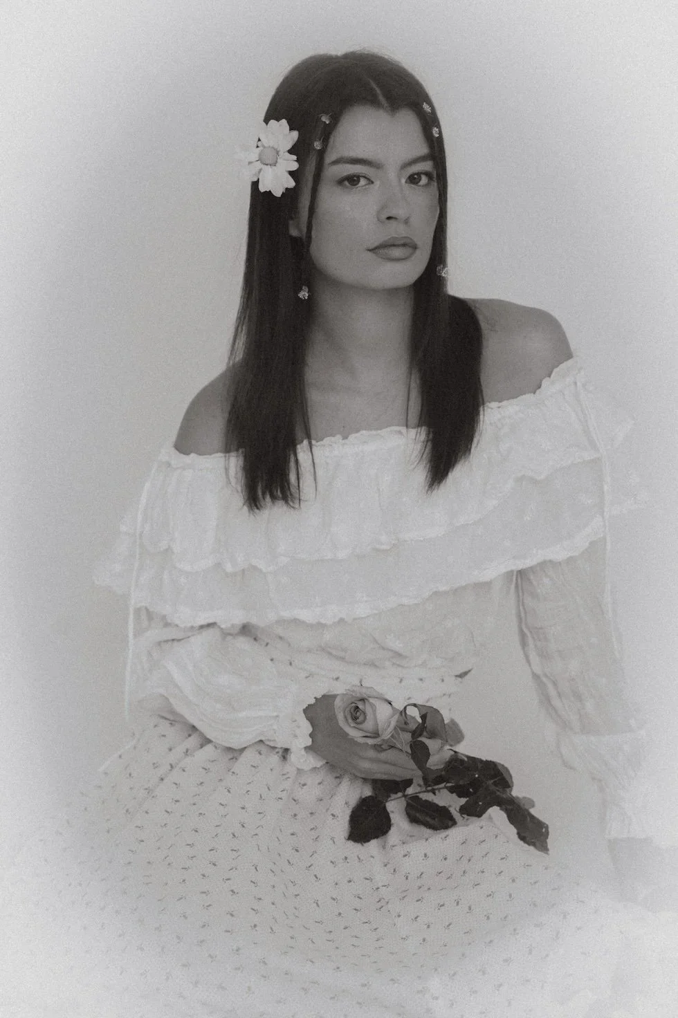 A woman with dark hair adorned with flowers, wearing an off-shoulder ruffled white dress, sitting and holding a single rose in her lap.