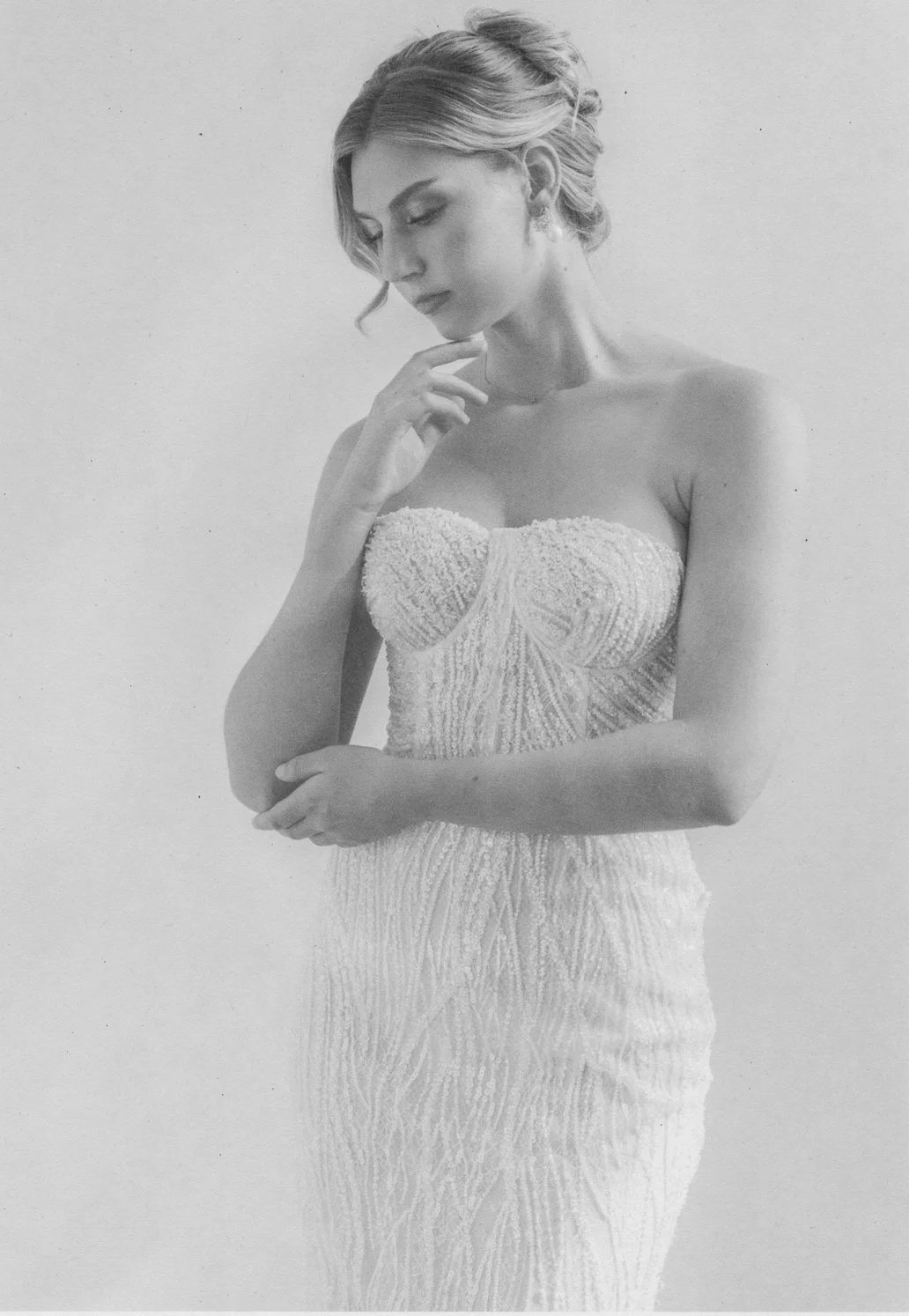 A woman in a strapless, textured, elegant dress with her eyes closed and a pensive expression, touching her chin with her right hand.