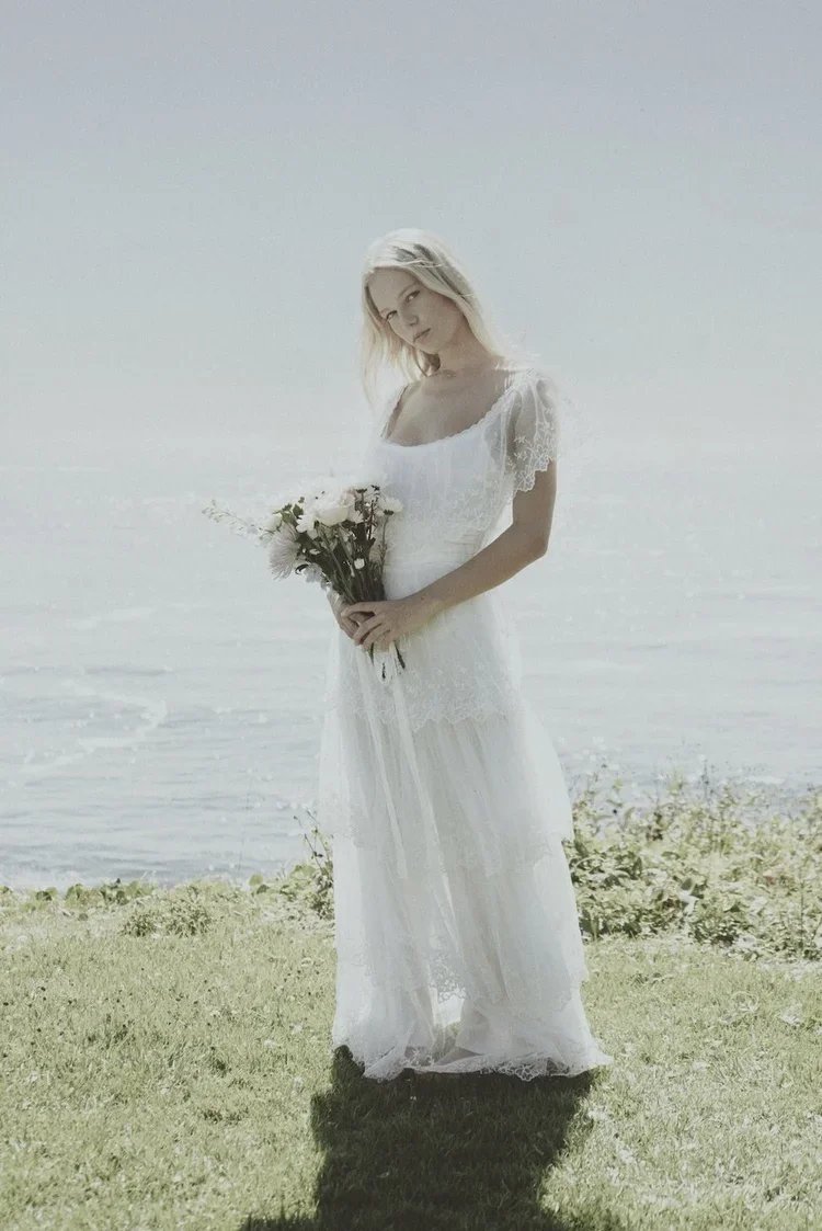 A woman in a white lace dress holding a bouquet of white flowers standing near a body of water on a grassy area.