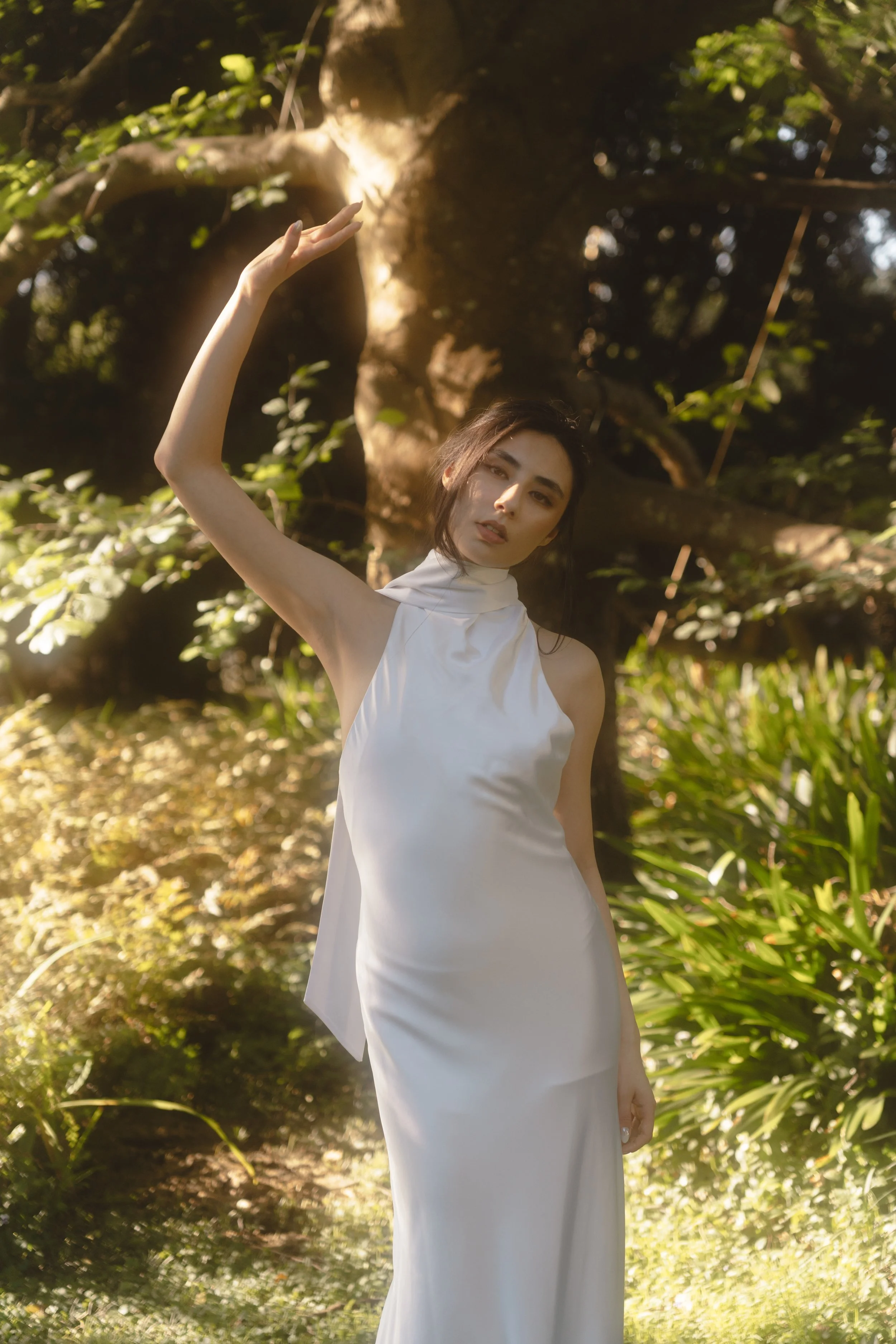 A woman in a white dress standing outdoors near a large tree with sunlight shining through the leaves.