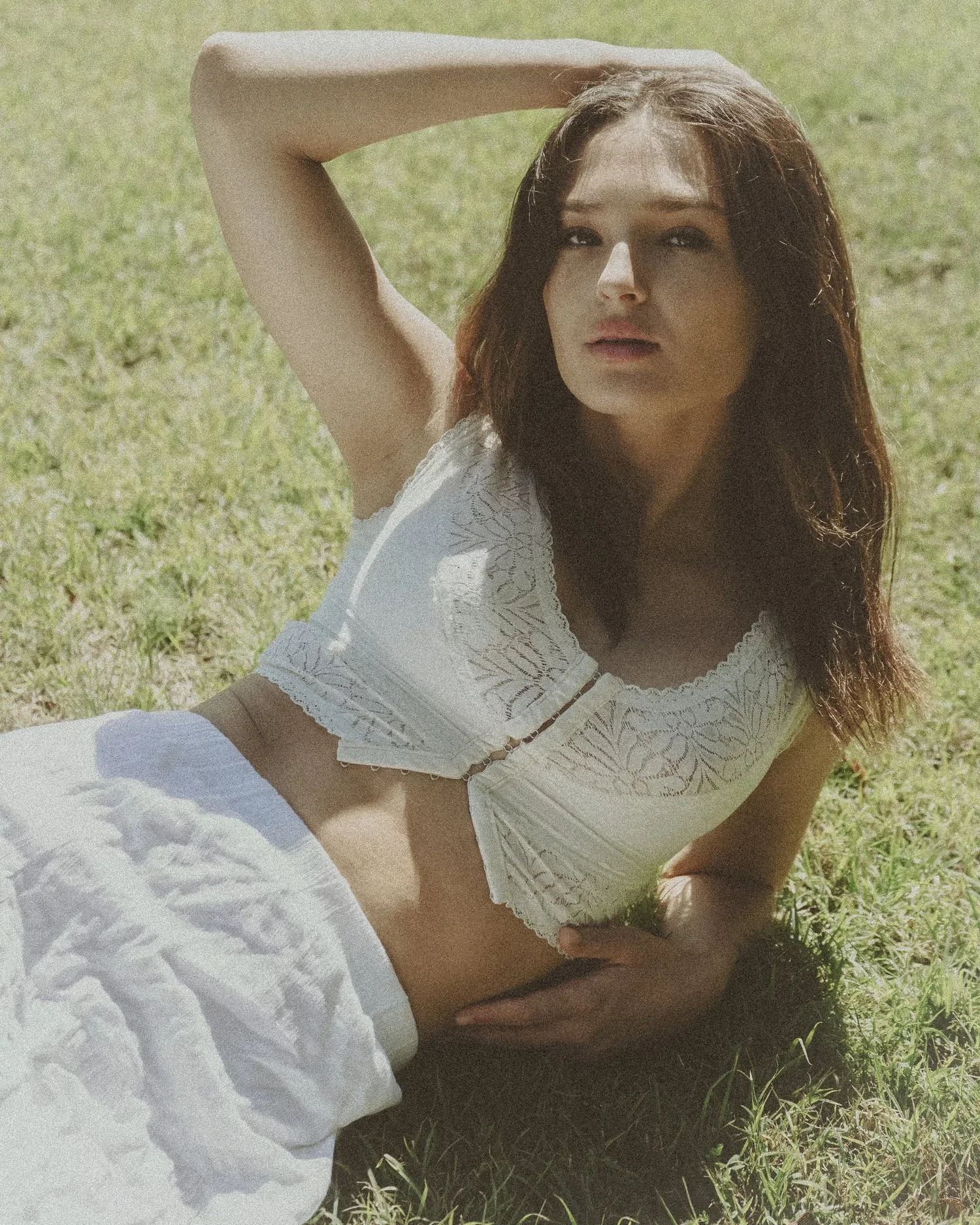 A woman with shoulder-length brown hair lying on grass, wearing a white sleeveless lace top and white pants, with one arm behind her head and one hand resting on her waist, looking at the camera.