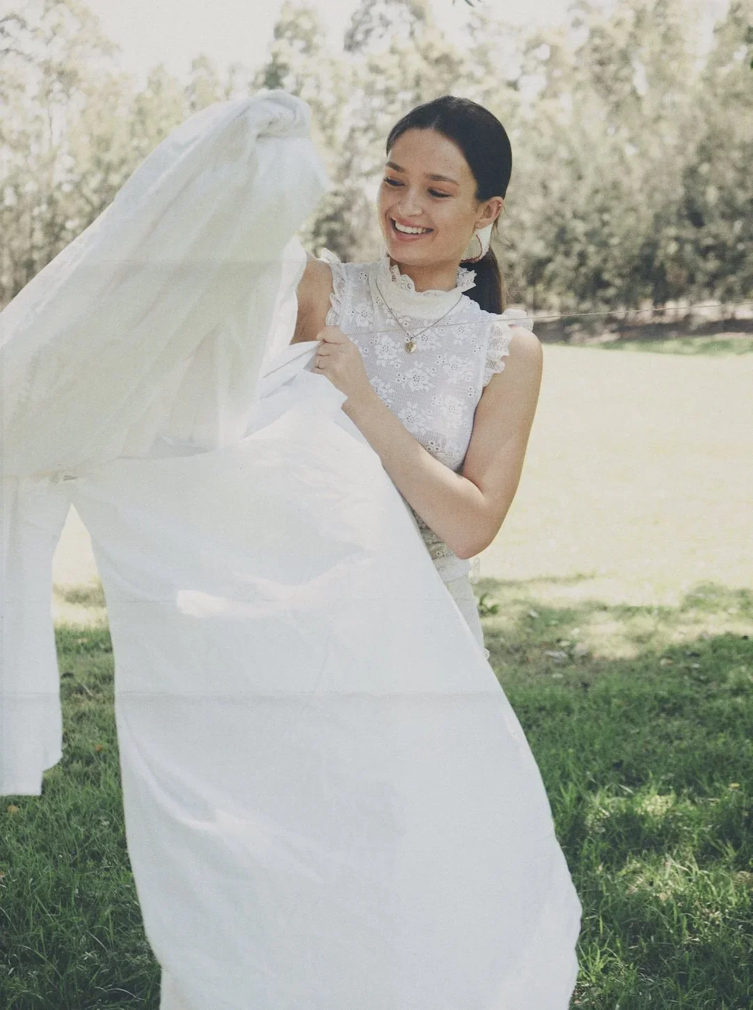 A young woman with dark hair wearing a white lacy sleeveless dress outdoors, smiling as she holds a white fabric or veil.
