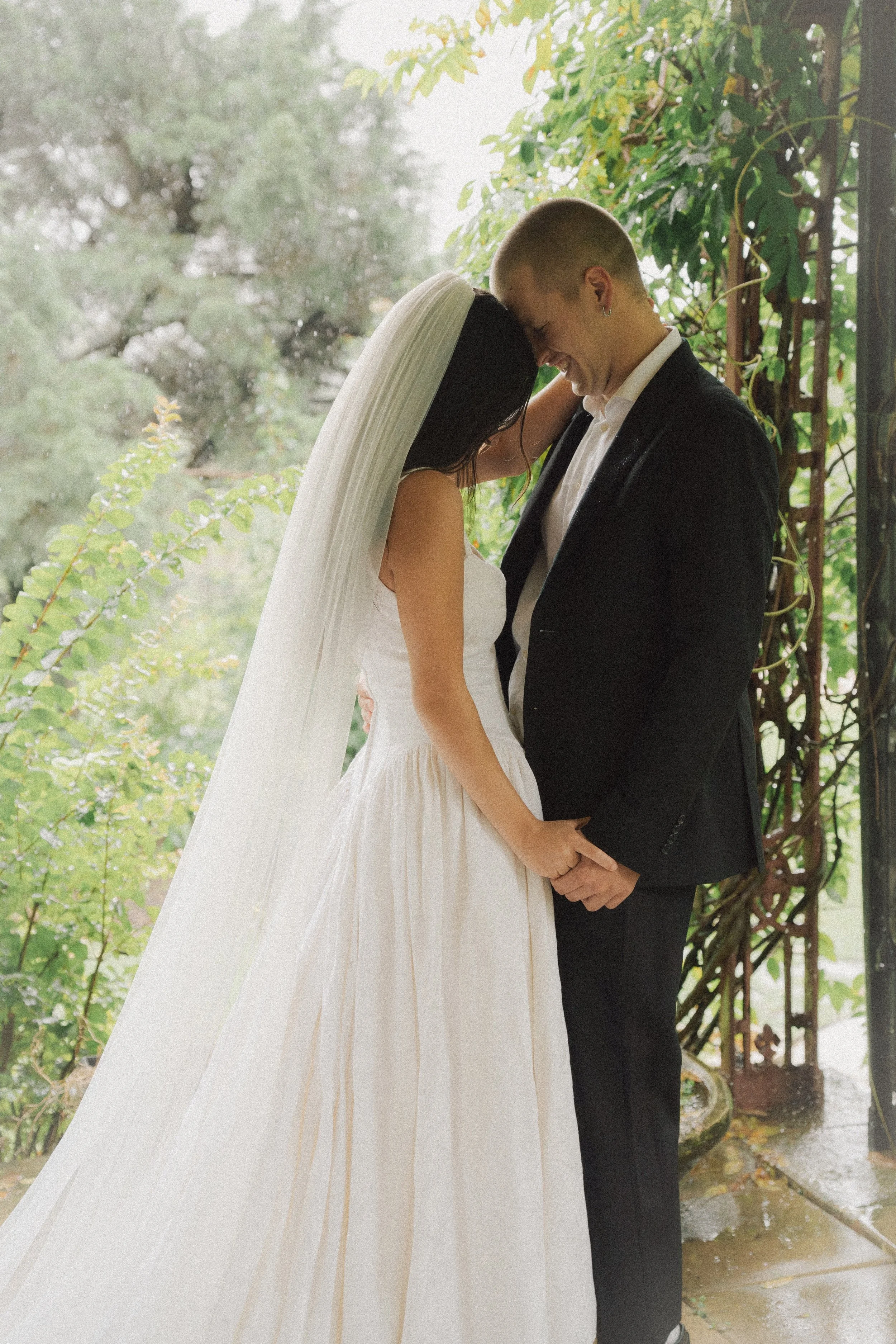 A bride and groom sharing a tender moment, smiling with foreheads touching, holding hands, standing under greenery with a romantic, natural backdrop.