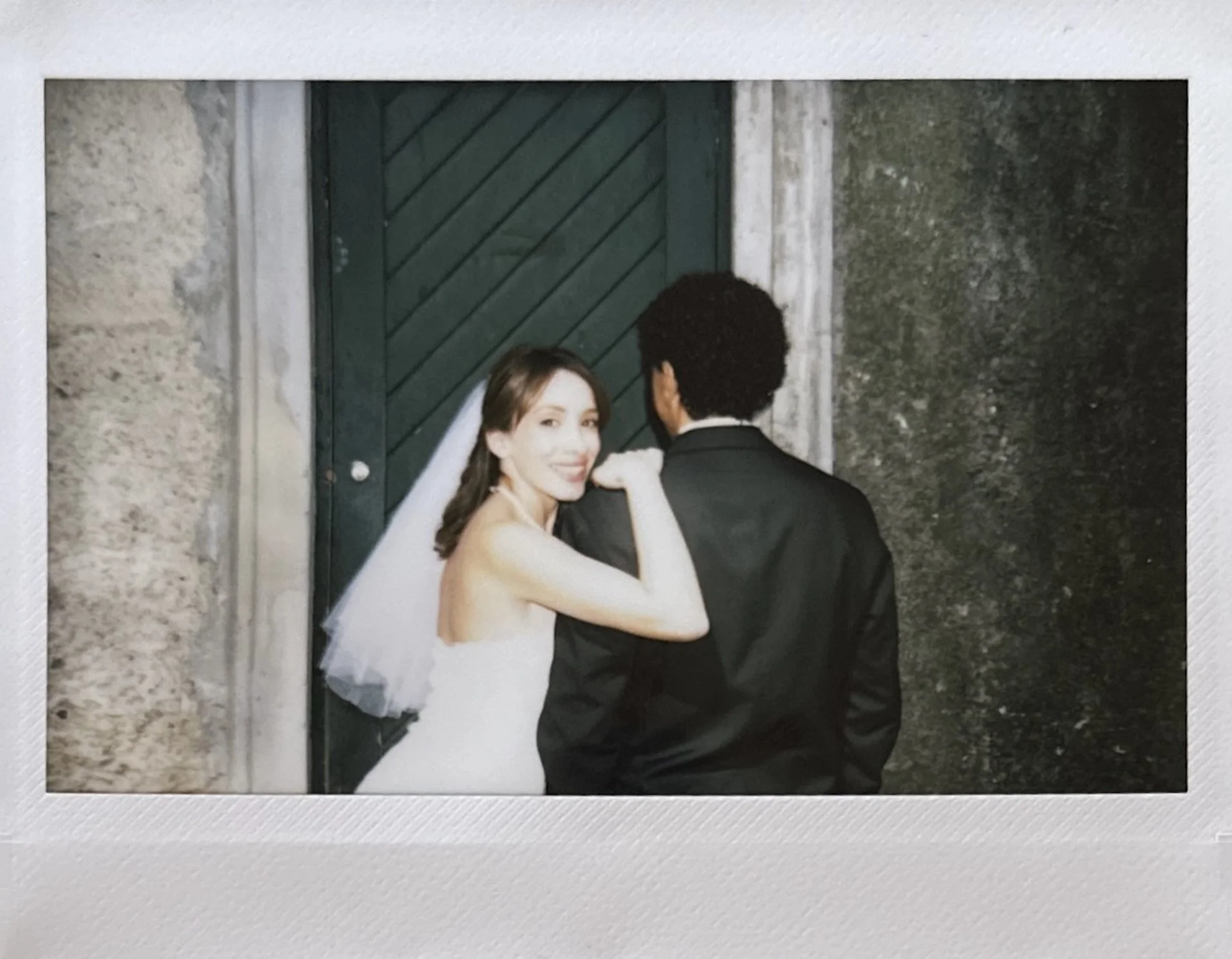 A bride with brown hair wearing a white wedding dress and veil, smiling and looking over her shoulder, with her arm resting on the back of a groom wearing a black suit. They are standing in front of a dark green door with a stone wall on the side.