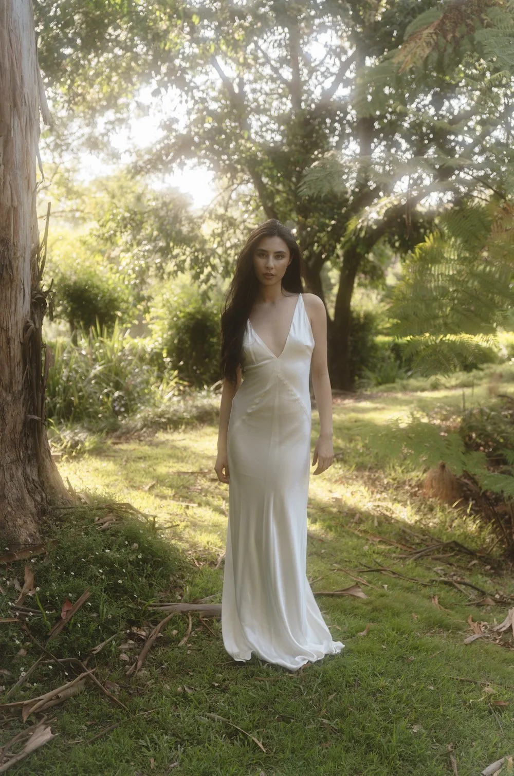 A woman in a white gown walking in a lush green garden with trees and sunlight.
