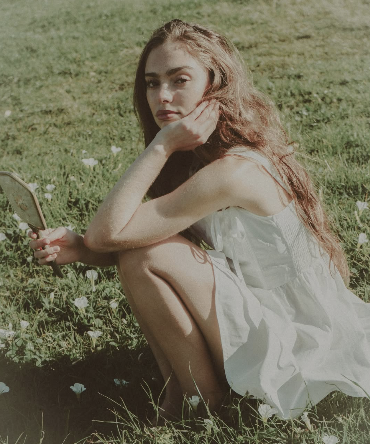 A young woman with long red hair sitting on grass in a white dress, holding a mirror and resting her face on her hand.