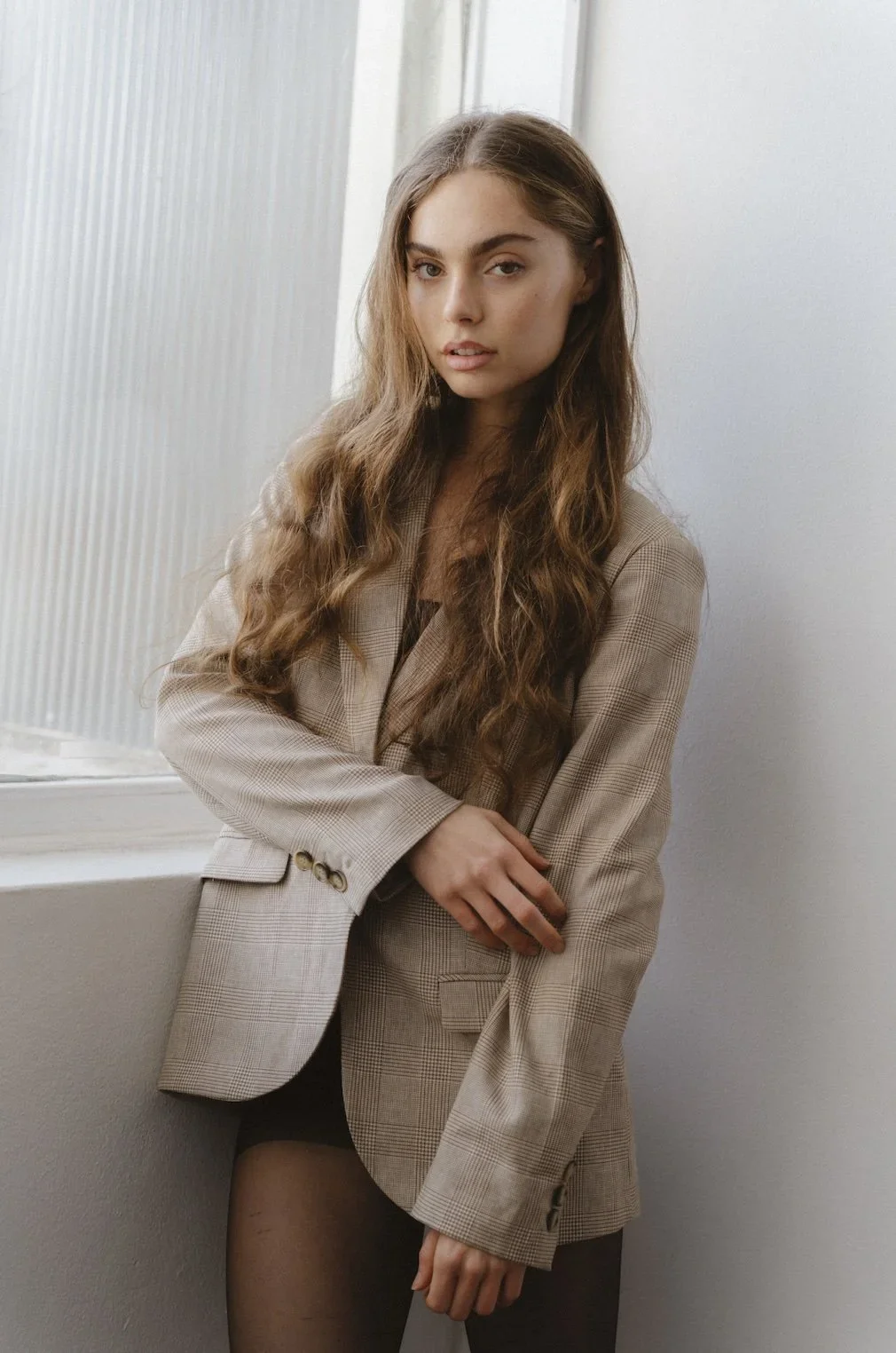 A young woman with long wavy hair wearing a beige checkered blazer and black tights, posing near a window with a neutral expression.