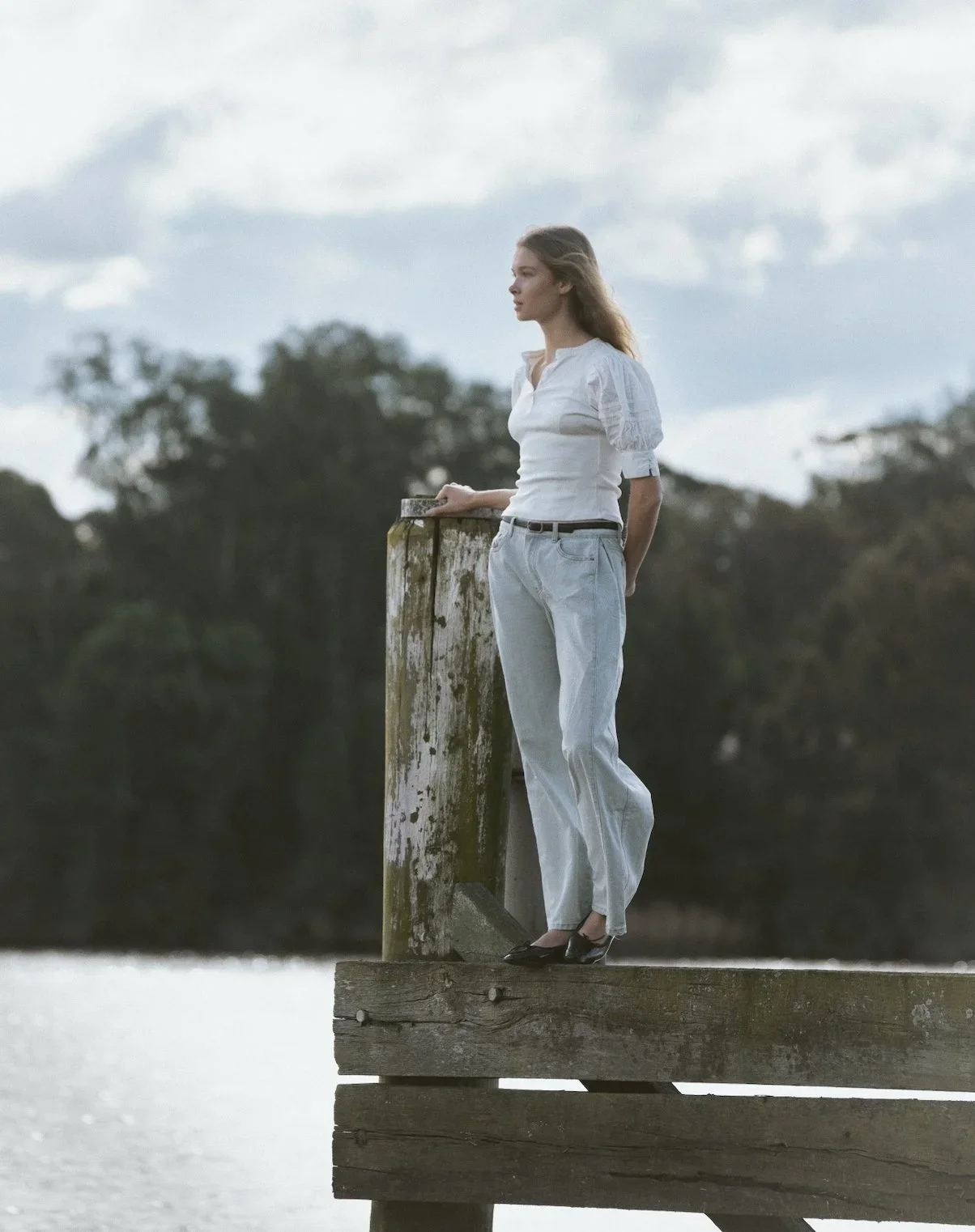 A woman standing on a wooden dock by a body of water, looking into the distance, with trees and a cloudy sky in the background.