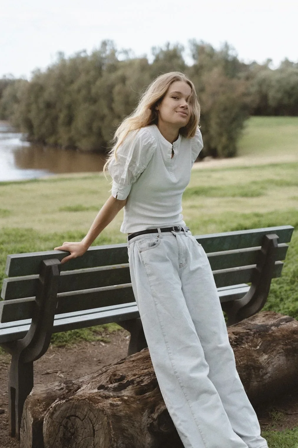 Young woman with blonde hair standing outdoors near a park bench, leaning against it with her hands, smiling slightly, with a river and trees in the background.