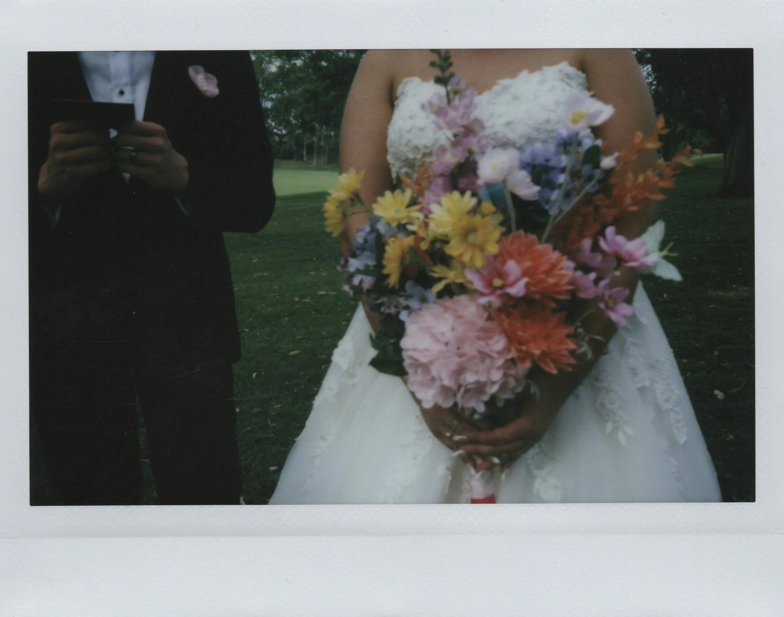 A bride holding a colorful bouquet of flowers and a man in a suit standing next to her in an outdoor park setting.