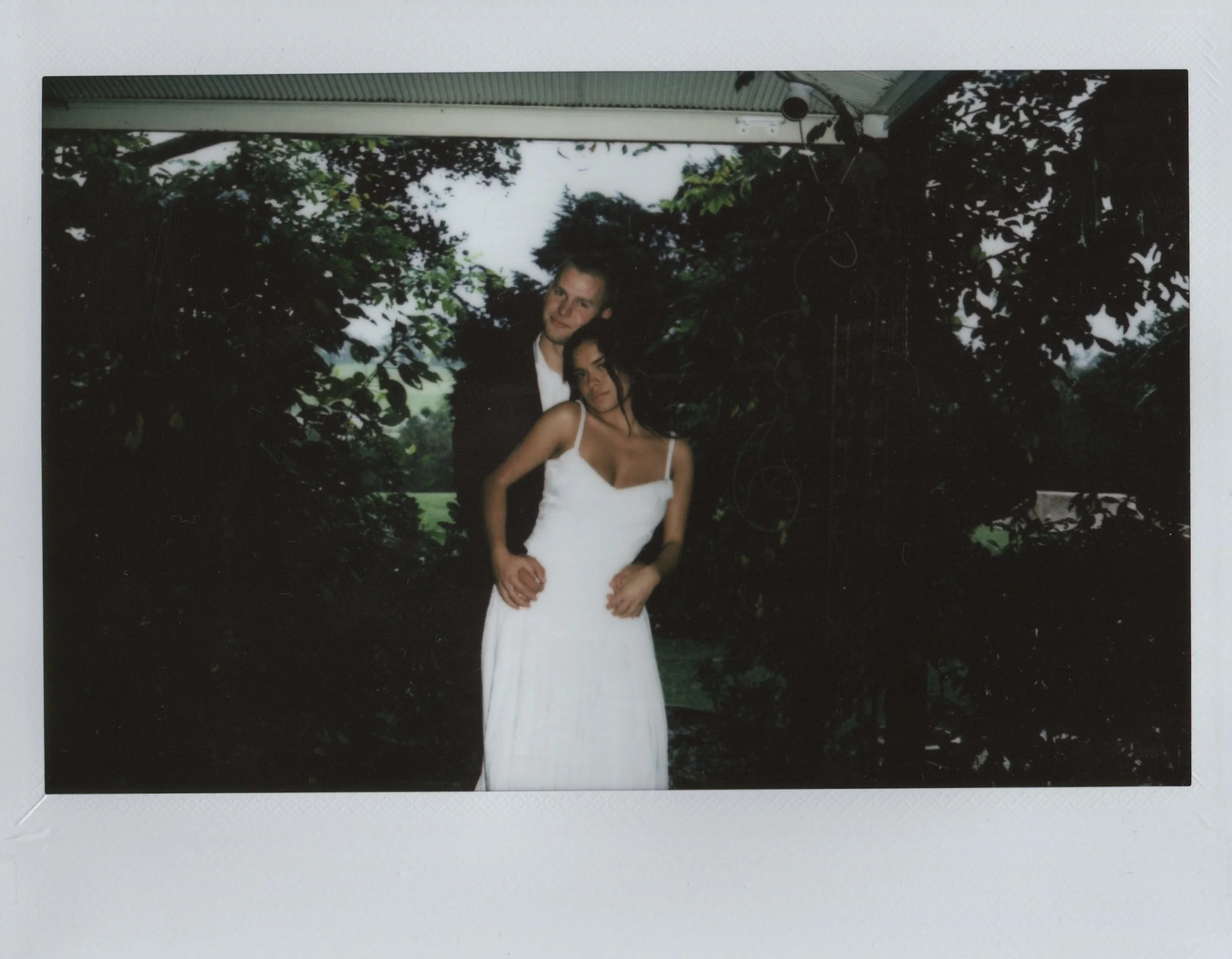 A man and woman in wedding attire posing together outdoors, surrounded by trees and greenery.