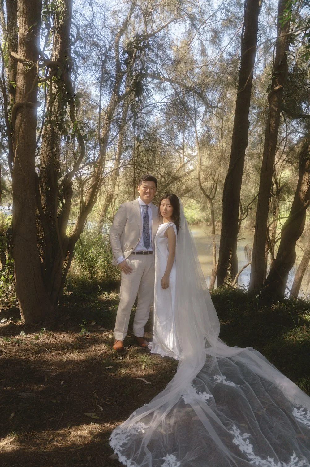 A bride and groom standing outdoors among trees beside a body of water. The bride is wearing a white wedding gown with a long lace train and veil, and the groom is dressed in a light-colored suit with a blue patterned tie. Both are smiling and lookin