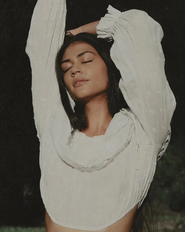 A woman with long dark hair and tan skin relaxing outdoors with eyes closed, wearing a loose white blouse with puffed sleeves and ruffles, standing against a dark background.