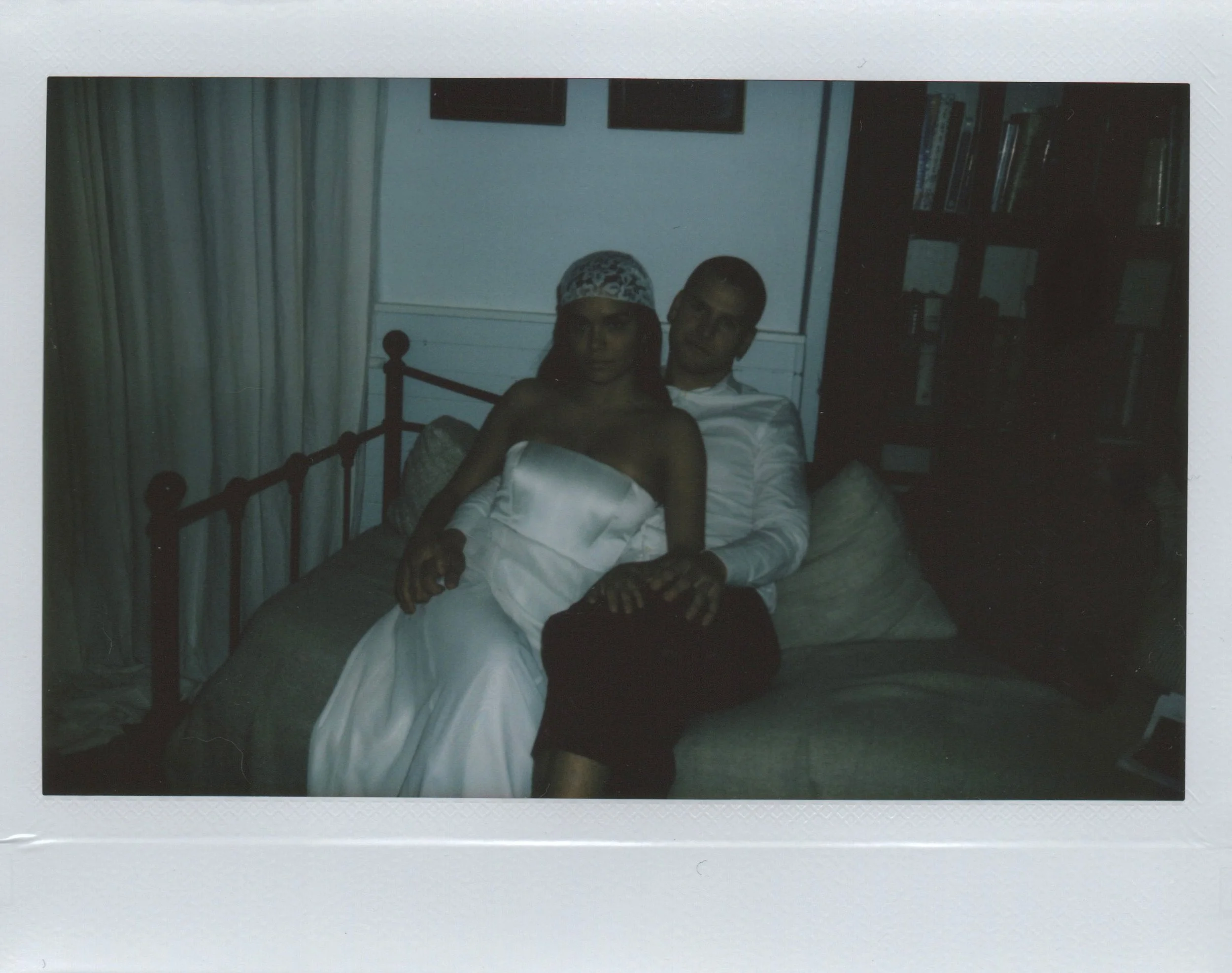 Two people sitting on a bed in a dimly lit room, one wearing a dress and headscarf, the other in a shirt and dark pants, with a shelf of books behind them.