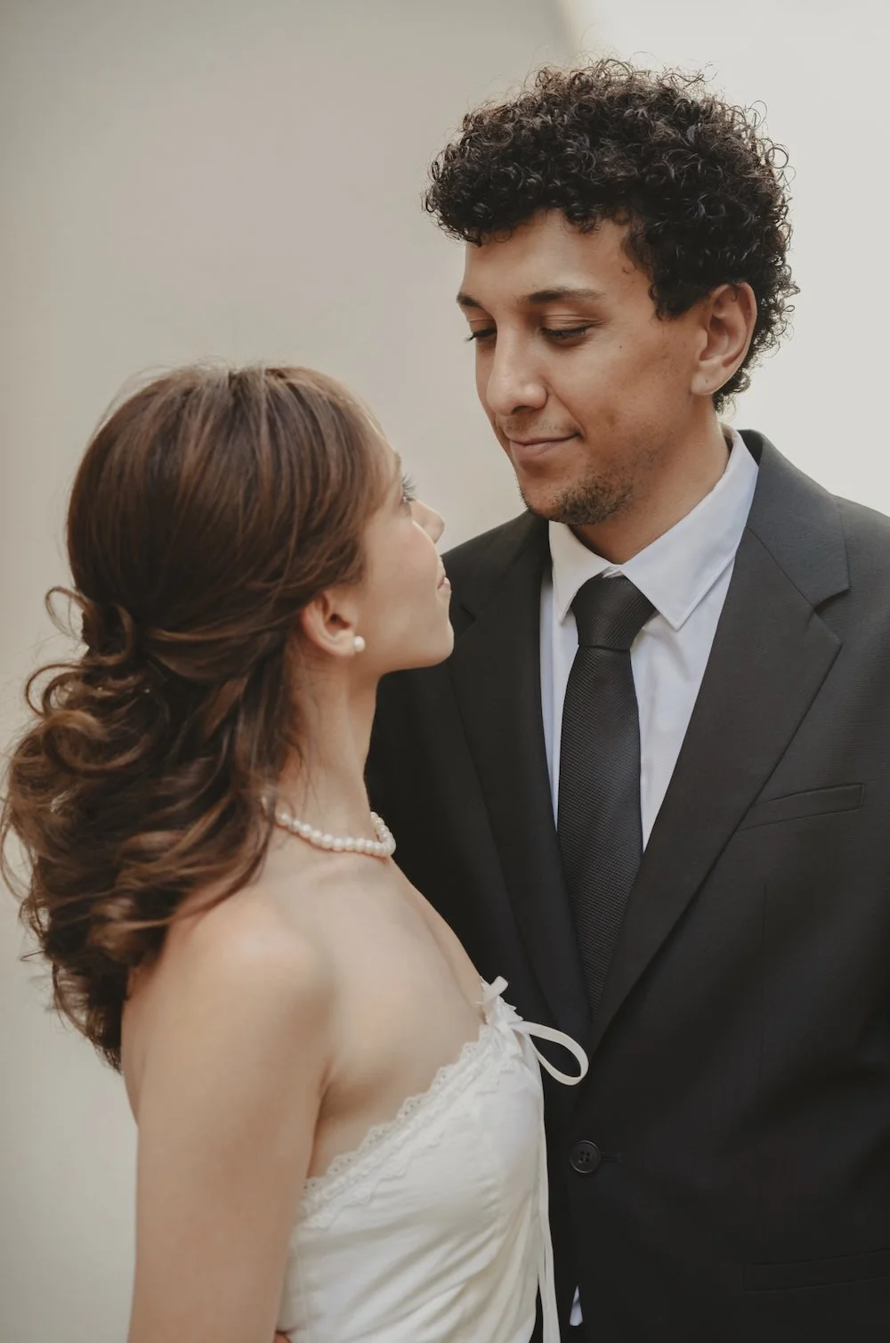 A bride and groom looking into each other's eyes at their wedding, with the bride wearing a pearl necklace and strapless white dress and the groom in a black suit and tie.