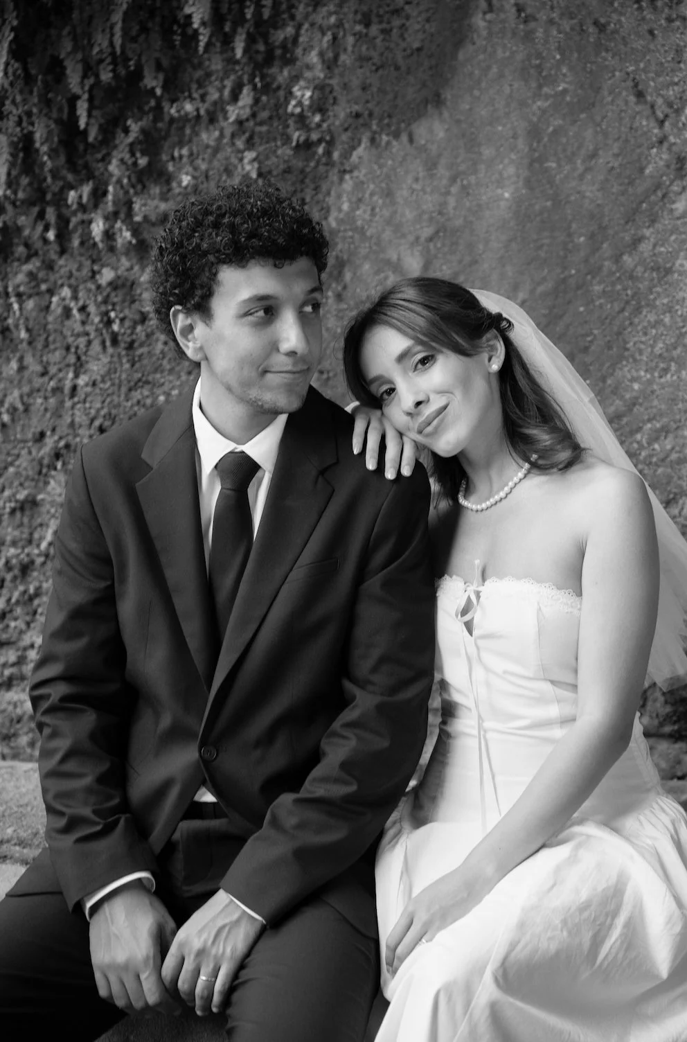 A black-and-white photo of a bride and groom sitting together outdoors. The groom, wearing a suit and tie, has curly hair and looks at the bride. The bride, in a strapless wedding dress with a pearl necklace and veil, leans on his shoulder and smiles
