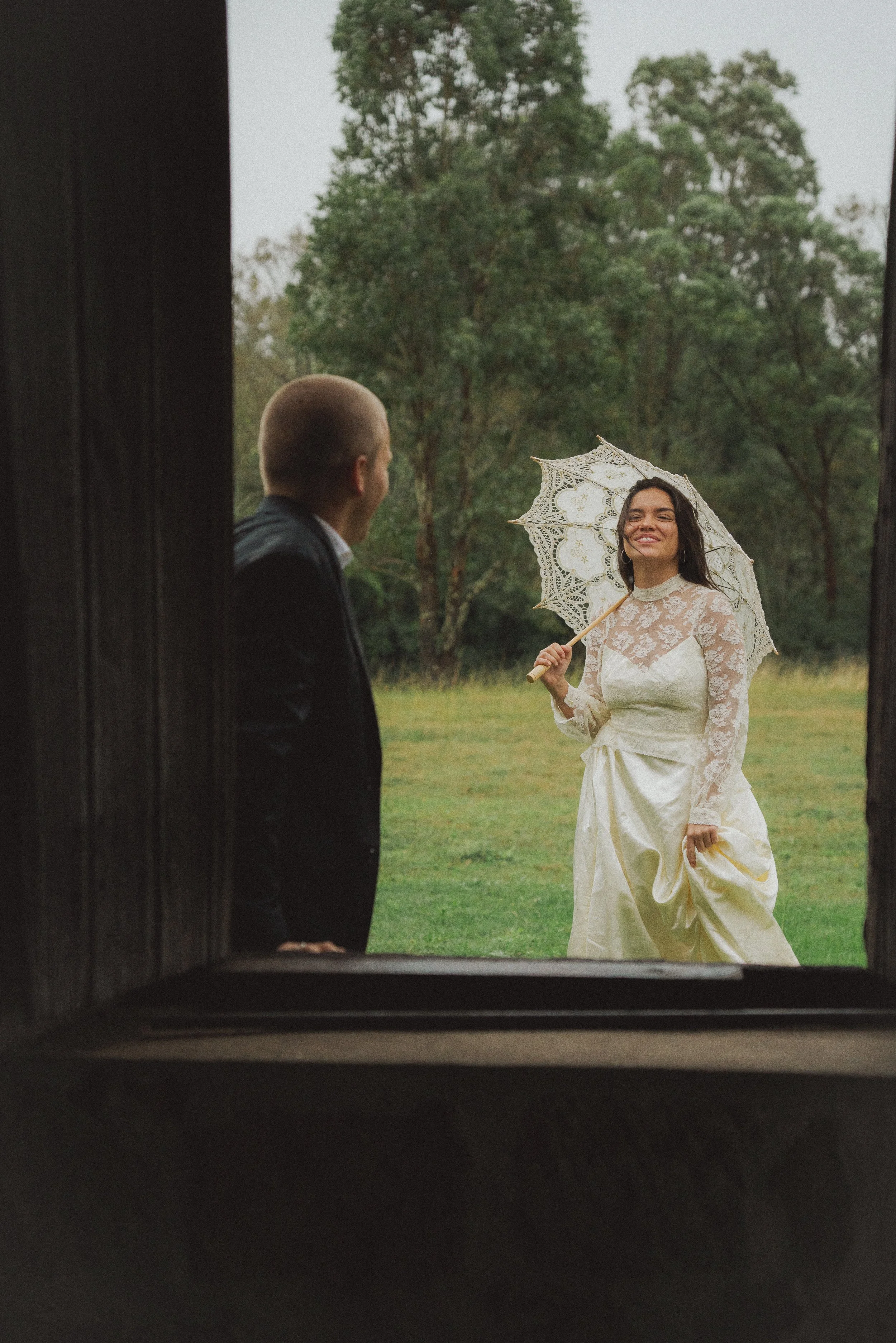 A bride in a white lace wedding dress holding a lace umbrella smiling at a groom in a black suit, seen through a window on a rainy day with trees in the background.