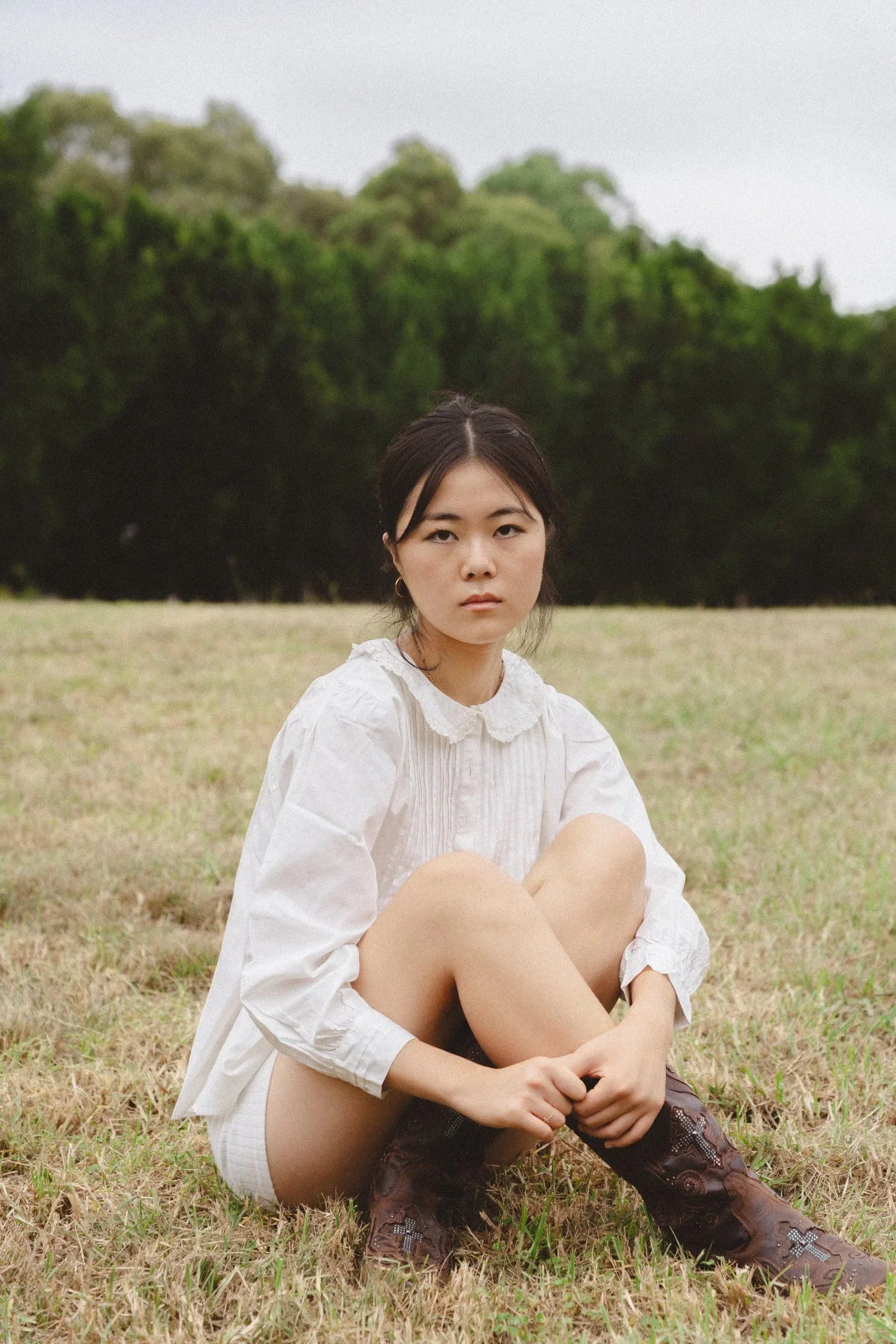 A young woman with dark hair sitting on grass in a field, wearing a white blouse and cowboy boots, with a forest in the background.