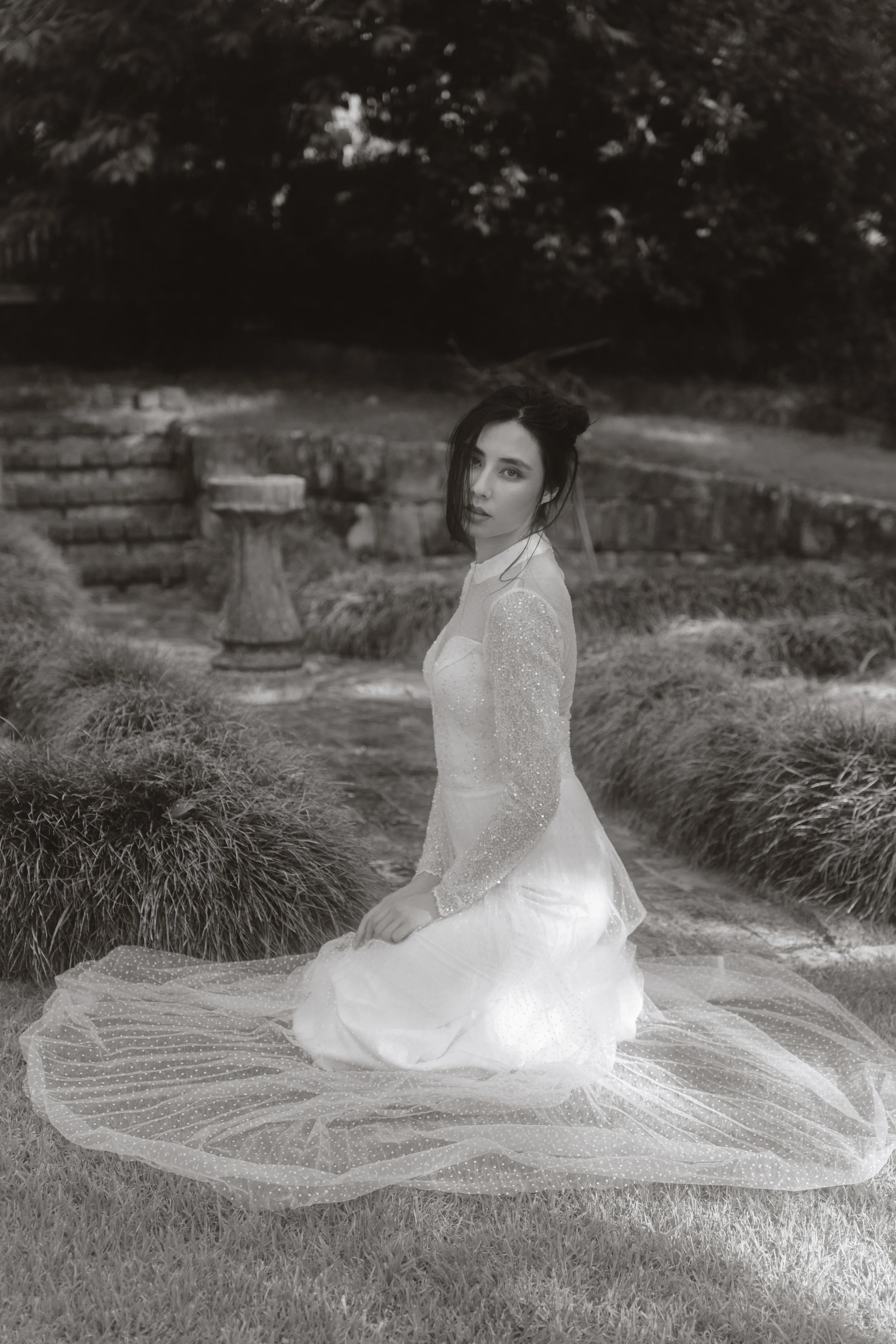 A woman dressed in a long, shimmering wedding gown sitting on the grass in a garden, with a stone pathway and lush trees in the background, captured in black and white.