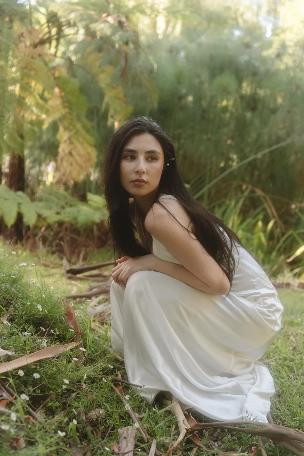 A young woman with long dark hair in a white dress crouches outdoors in a lush, green forest setting, surrounded by foliage and sunlight filtering through the trees.