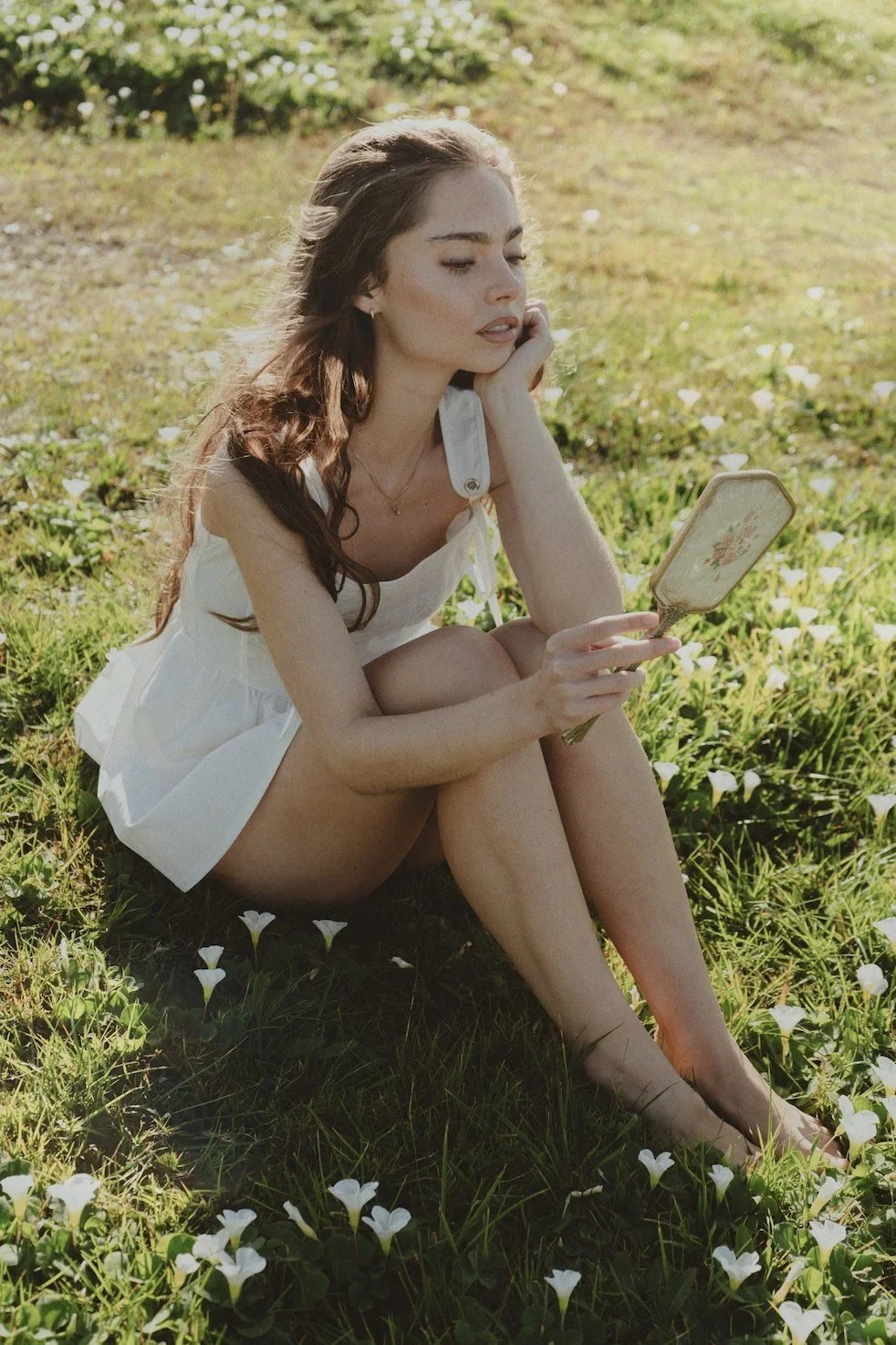 A young woman with long brown hair sitting barefoot on grass among white flowers, looking into a small floral mirror.