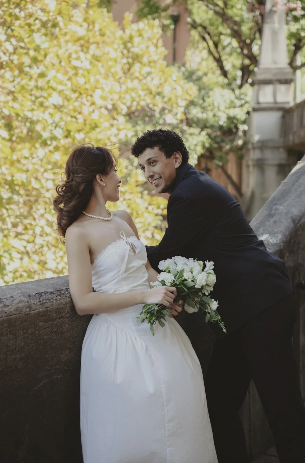 A bride and groom share a romantic moment outdoors during their wedding, with the bride holding a bouquet of white flowers and the background filled with green and yellow foliage.