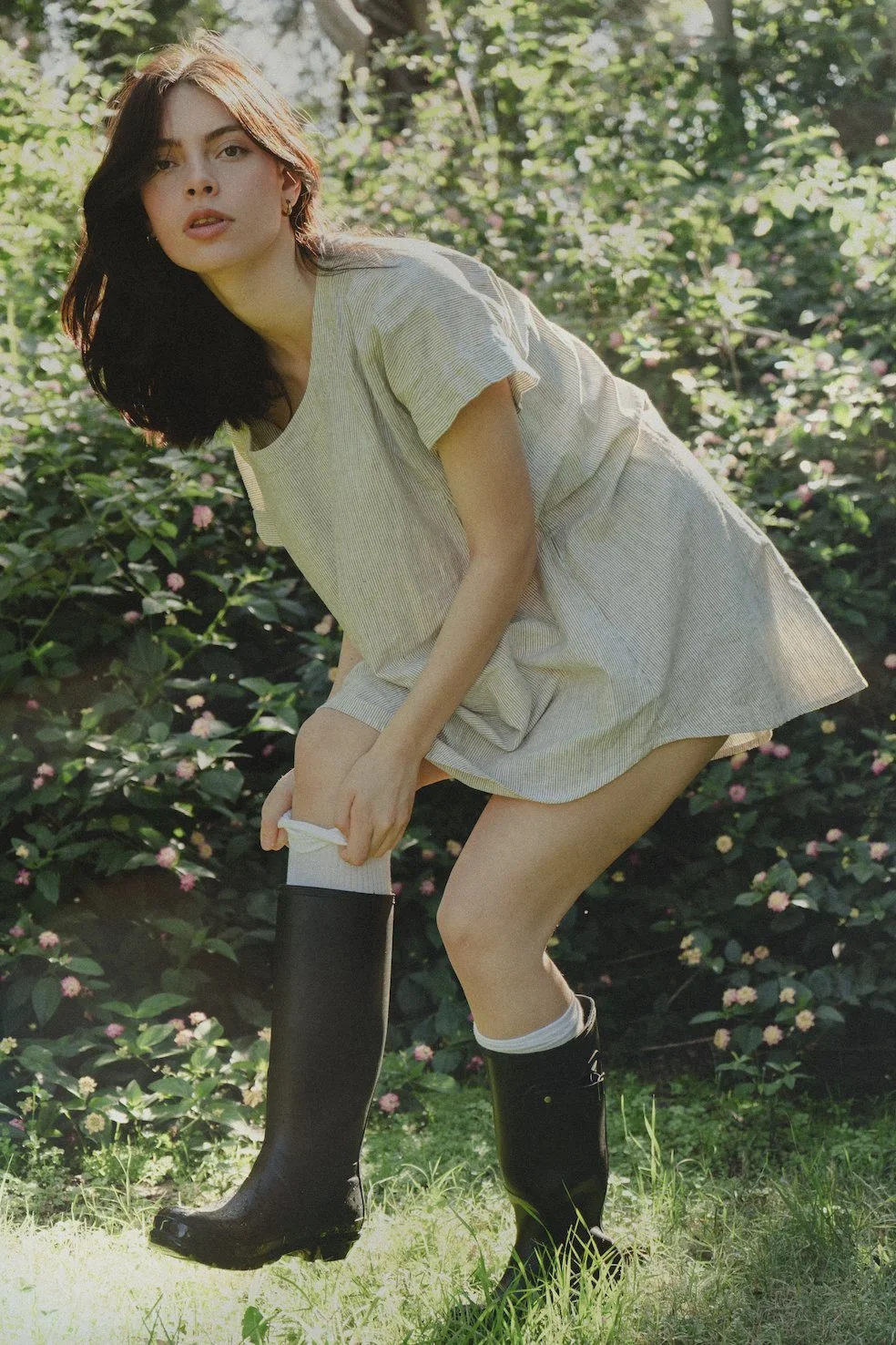 A young woman with dark hair wearing a beige dress and black rubber boots, kneeling outdoors in a garden with pink and white flowers.