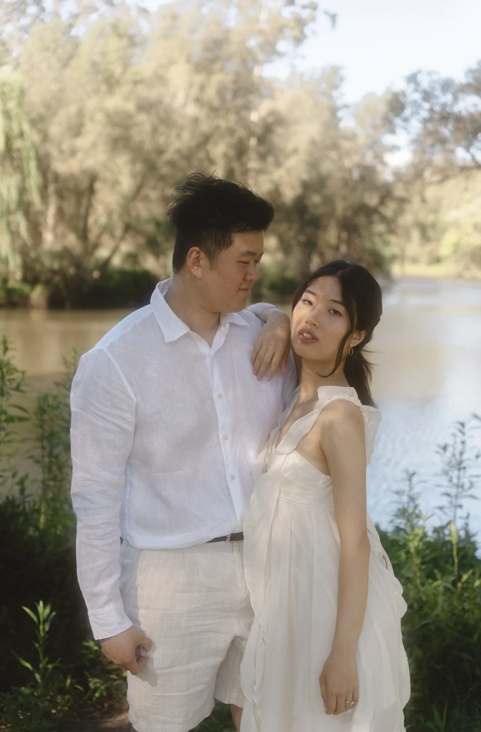 A couple standing outdoors near a river, with trees in the background, dressed in white and looking into each other's eyes.