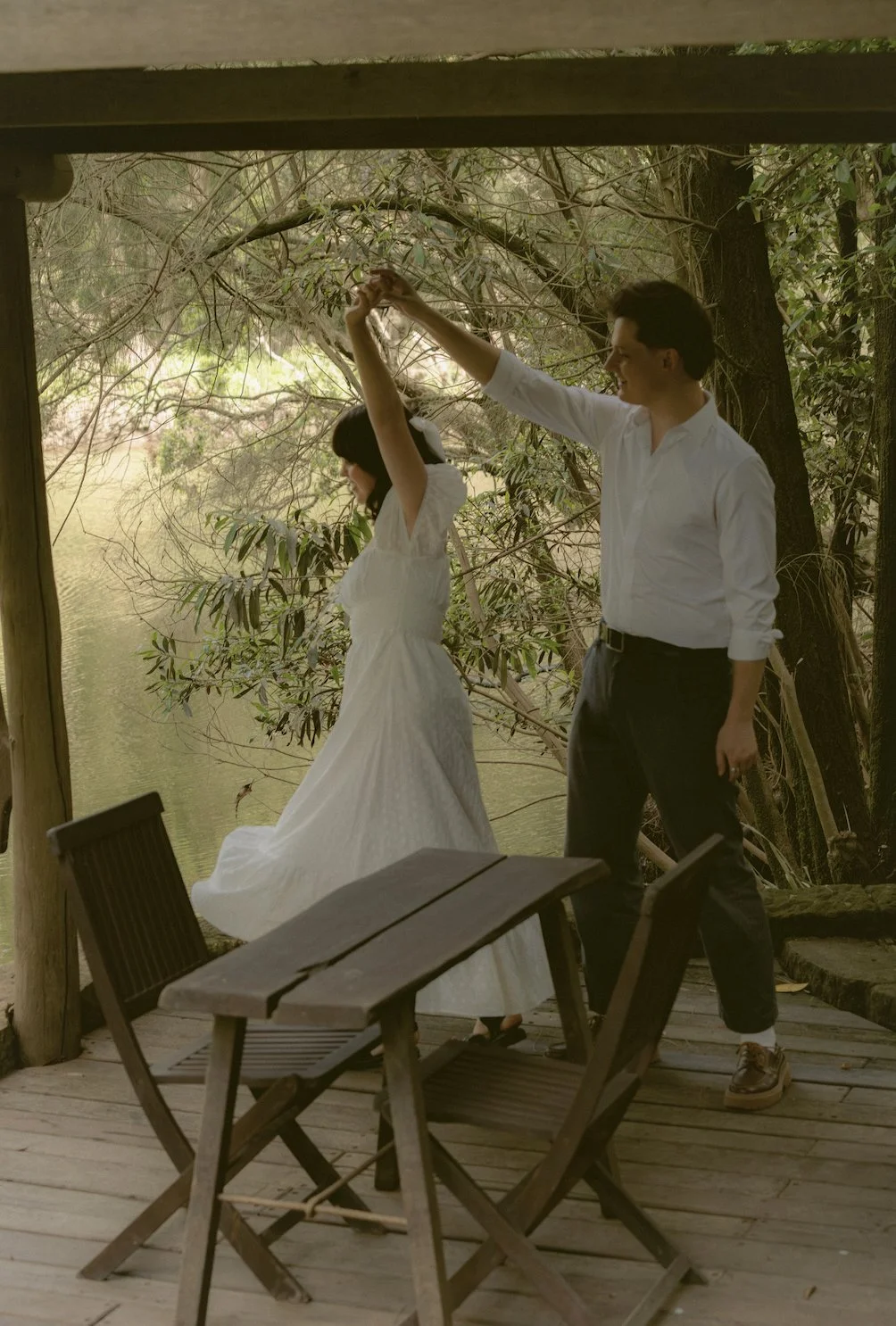 A man twirling a woman in a white dress near a pond surrounded by trees.