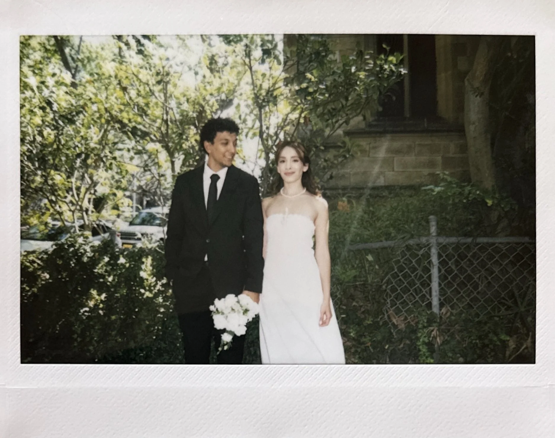 A couple in wedding attire standing outdoors on a sunny day, with the man in a black suit and holding a bouquet, and the woman in a white wedding dress, surrounded by greenery and a stone building.