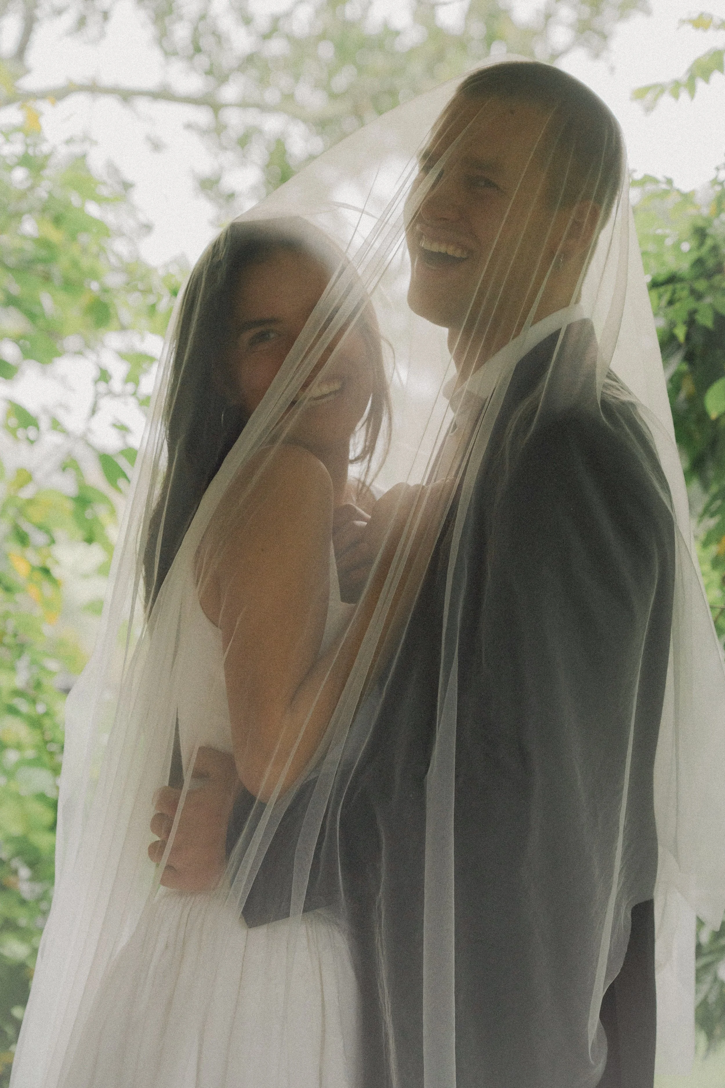 A smiling couple standing behind a sheer curtain, with greenery in the background, the woman wearing a white dress and the man in a dark suit, both happily looking at each other.