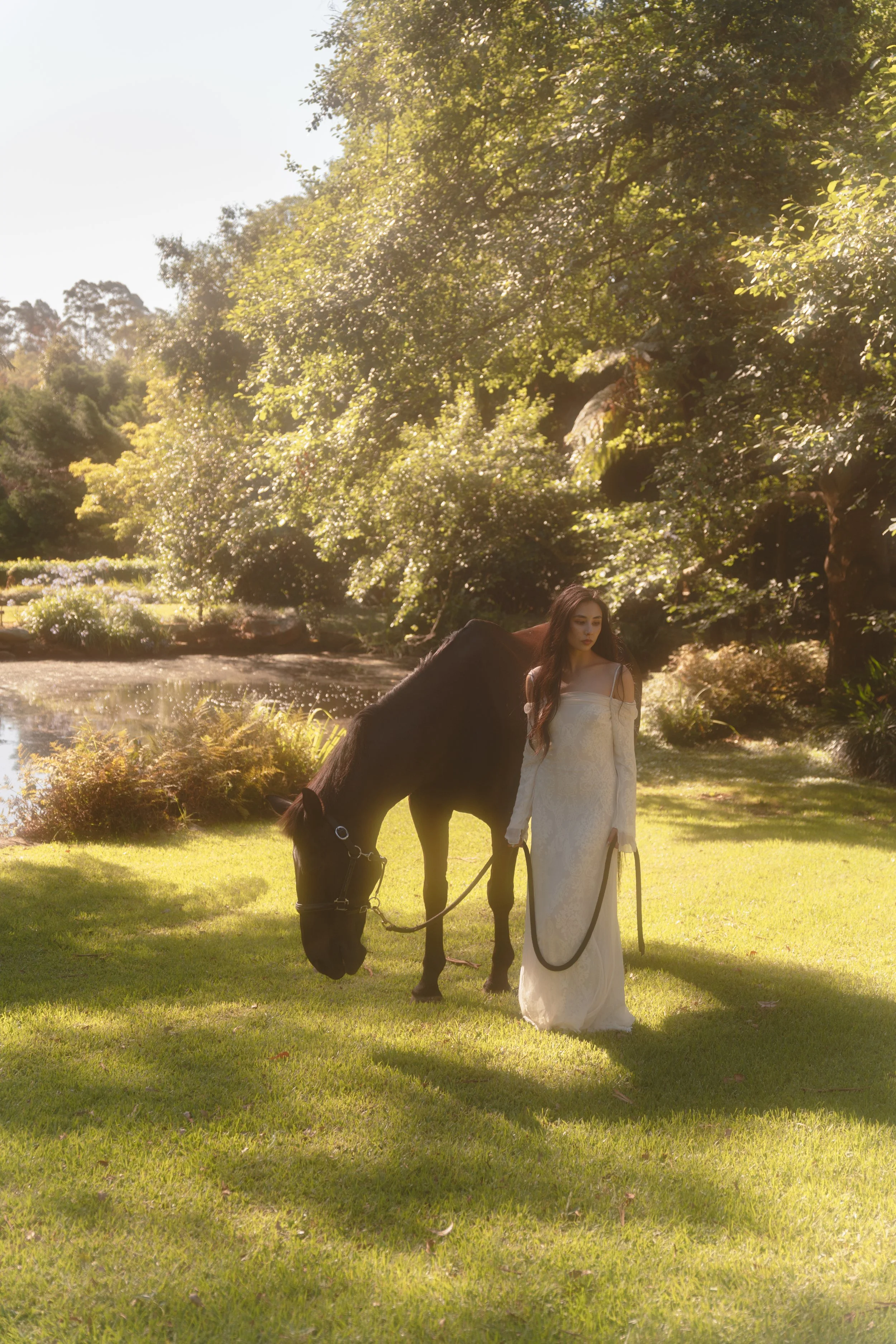 A woman in a white dress holding a black horse's reins stands on a grassy area near a pond with lush trees and bushes in the background on a sunny day.