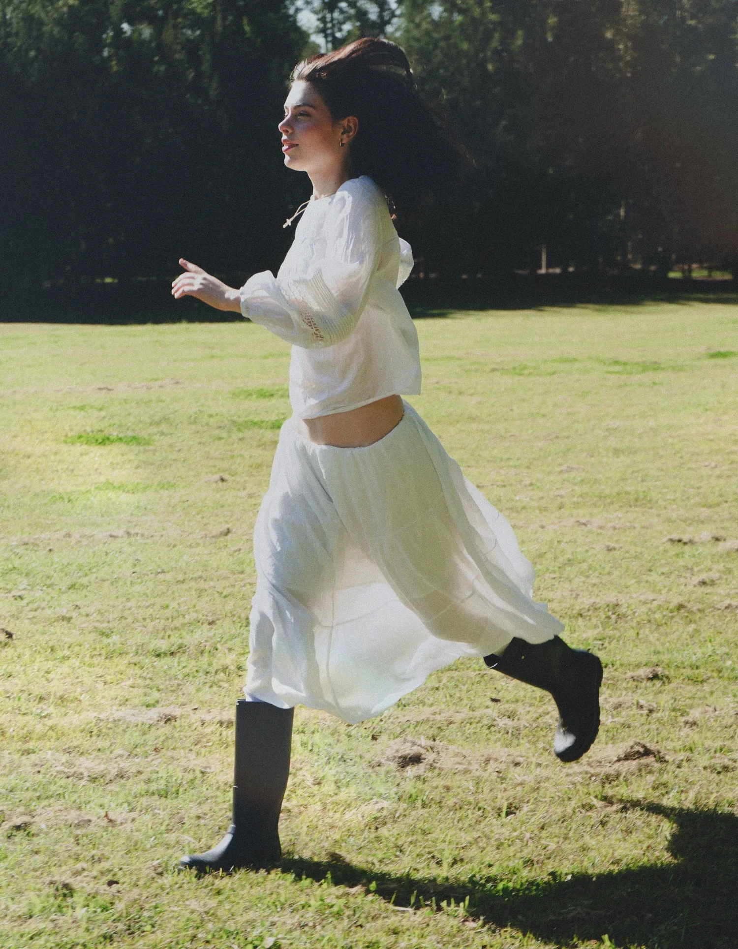 A woman in a white long-sleeve blouse, white skirt, and black rain boots runs across a grassy field with trees in the background.