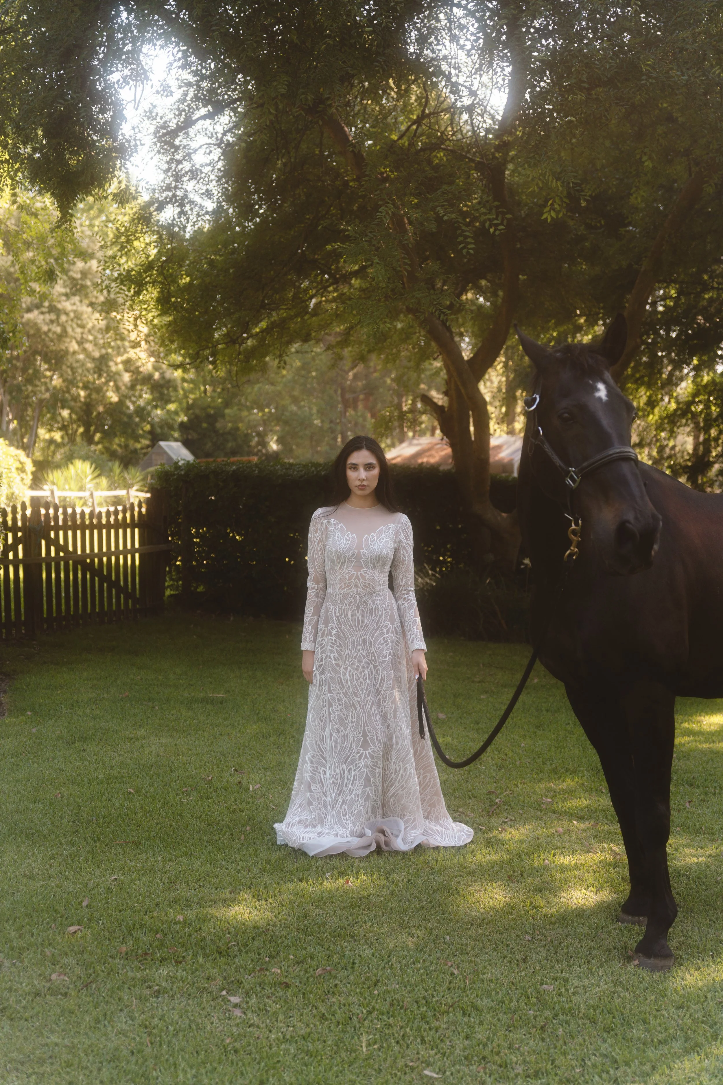 A woman in a long, white lace dress standing on a grassy lawn next to a black horse in a backyard with trees and a wooden fence.