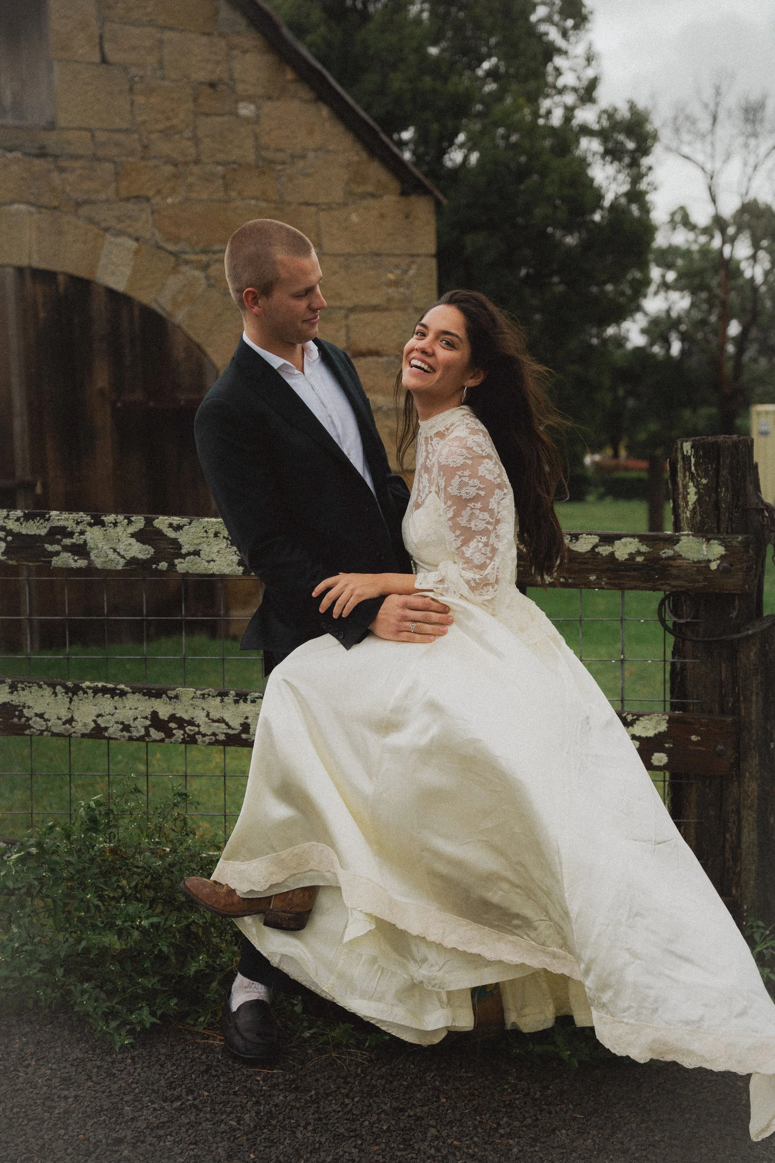 A smiling bride and groom in wedding attire outdoors near a rustic wooden fence and stone building.