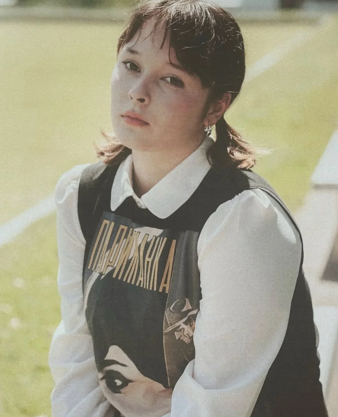 A young woman with short brown hair and earrings, wearing a white shirt and a black graphic vest, sitting outdoors on a grassy field.