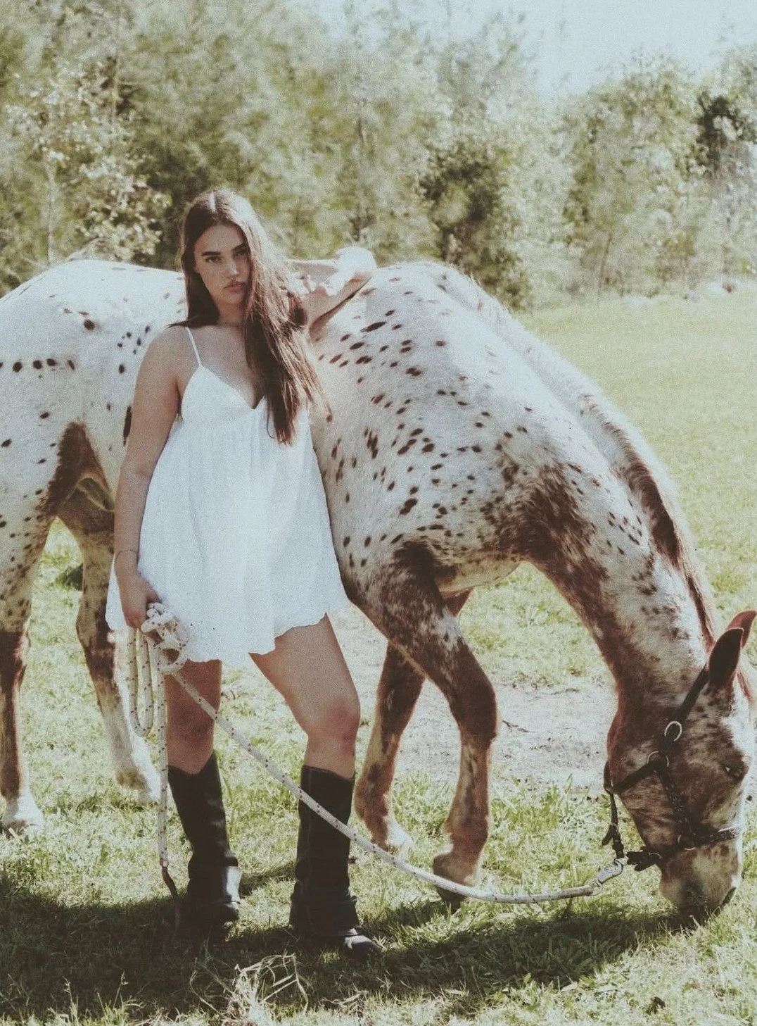 A young woman in a white dress and black boots standing next to a spotted horse grazing on grass outdoors.