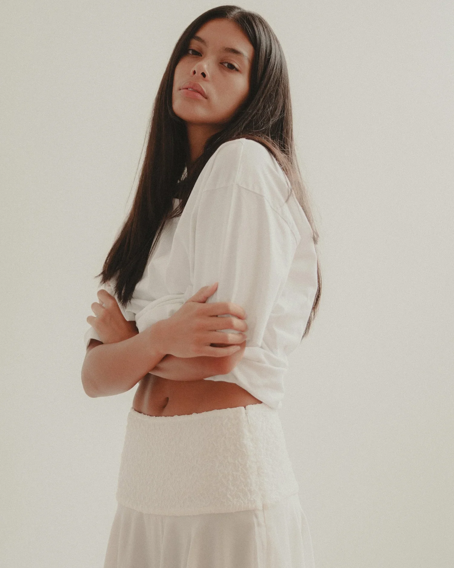 A young woman with long dark hair stands against a plain light-colored background, wearing a white top and skirt, with arms crossed and a neutral facial expression.