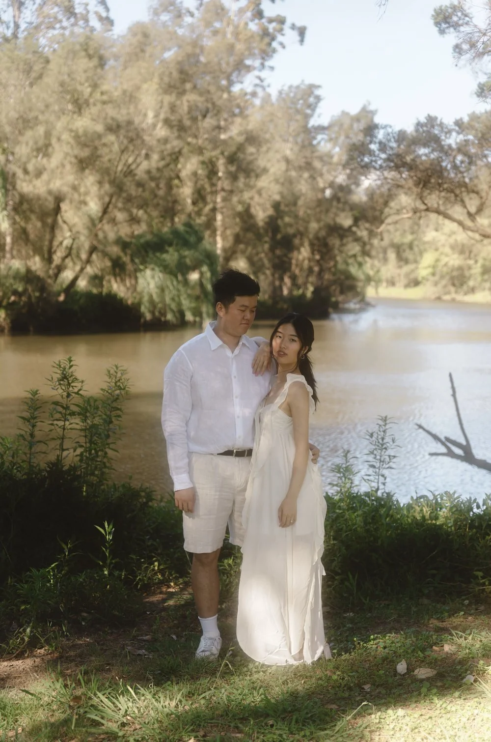 A young man and woman dressed in white stand by a river in a natural setting with trees in the background. The man is looking down at the woman with his arm around her shoulder, and the woman has her hand on her hip, gazing at the camera.