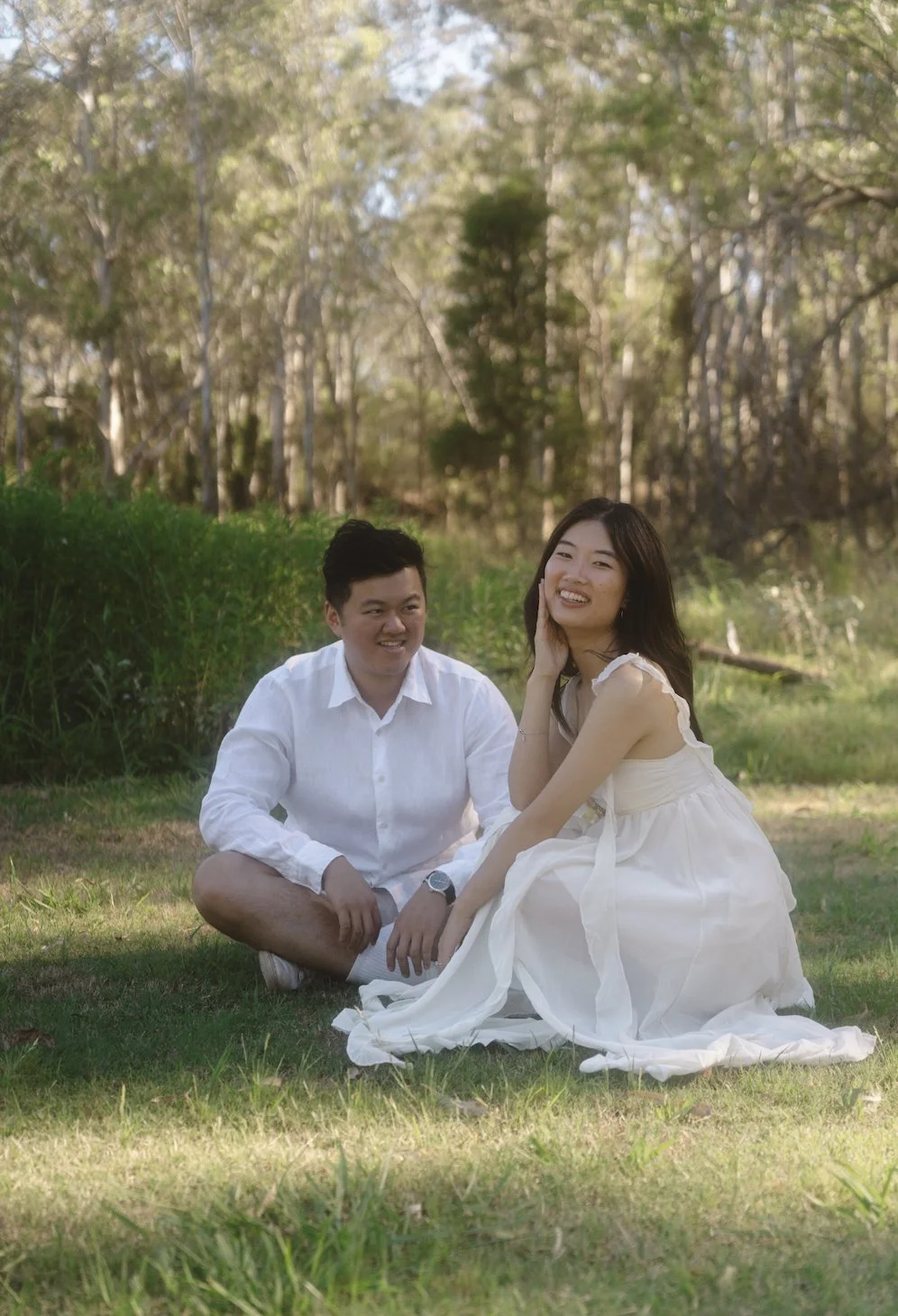A man and a woman sitting on grass in a wooded area, smiling and enjoying a relaxed moment.