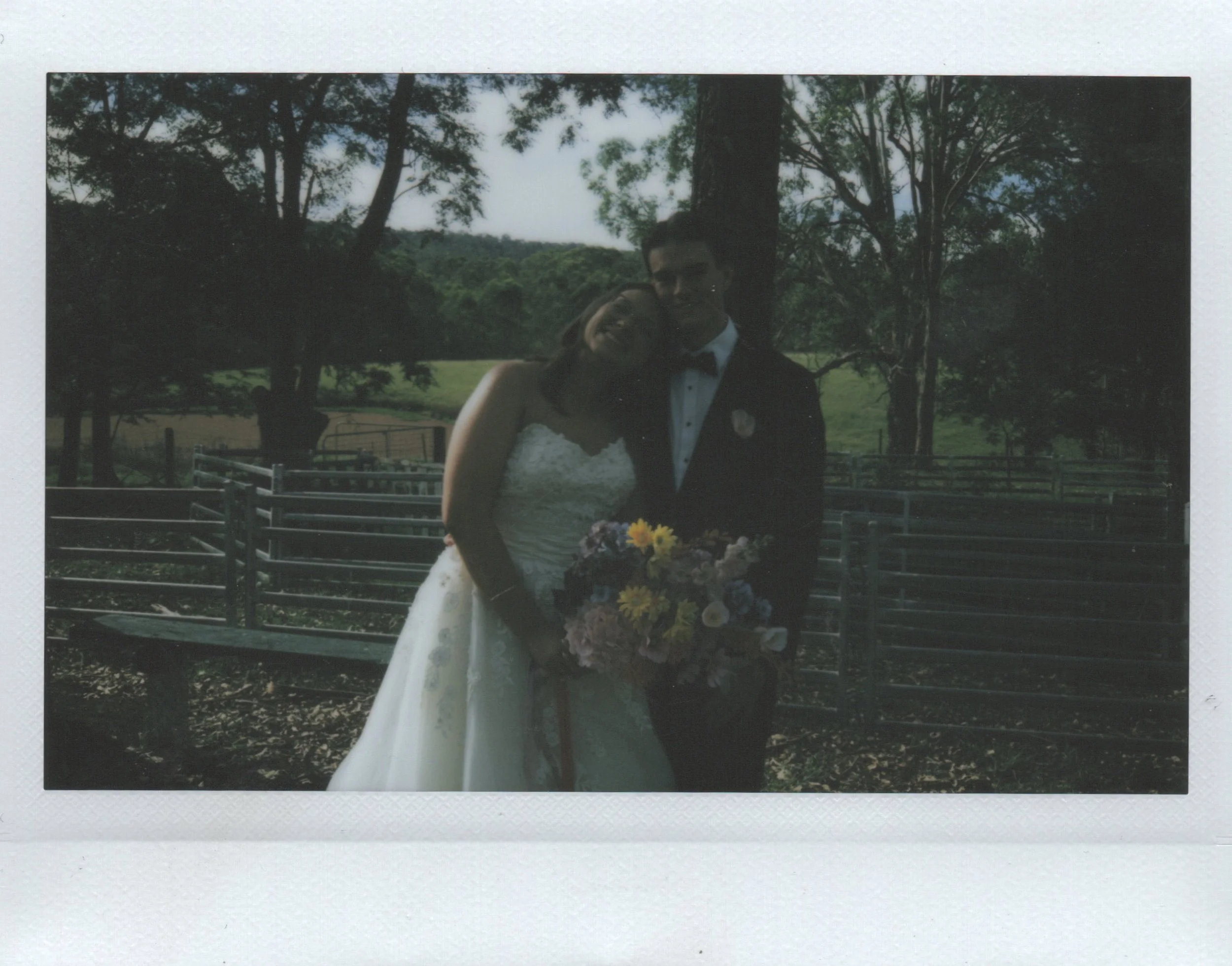 A bride and groom smiling and hugging outdoors during a wedding, with trees and a wooden fence in the background. The bride is holding a bouquet of flowers.