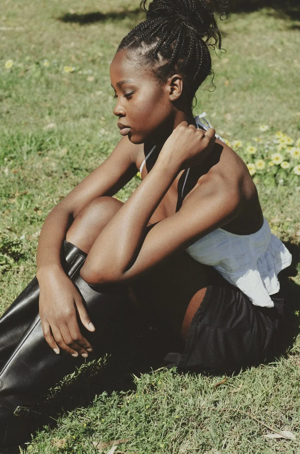 A young woman with braided hair sits on the grass, resting her chin on her hand, surrounded by small white and yellow flowers.