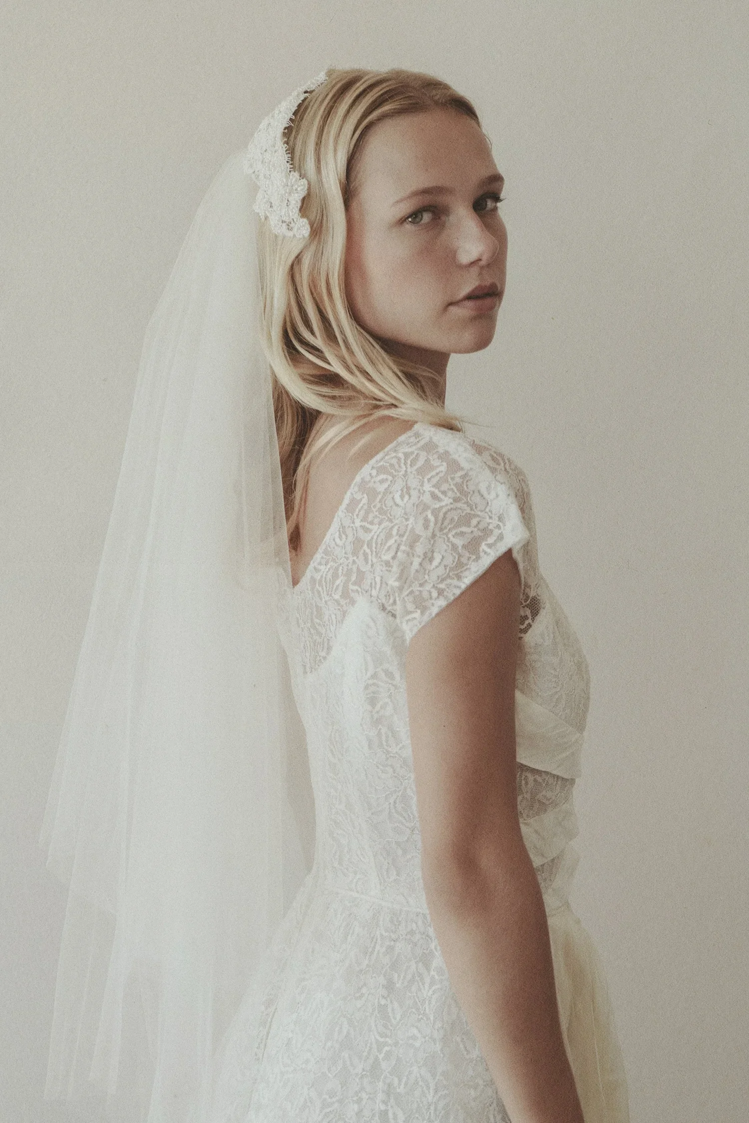 A young woman wearing a white lace wedding dress and a veil, looking over her shoulder against a plain background.