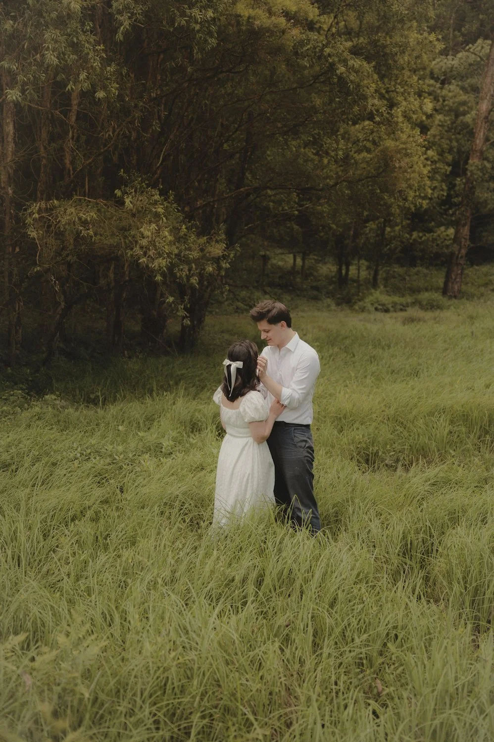 A young couple dressed in vintage clothing stands in a lush green field, holding hands and gazing at each other, surrounded by tall grass and trees in the background.