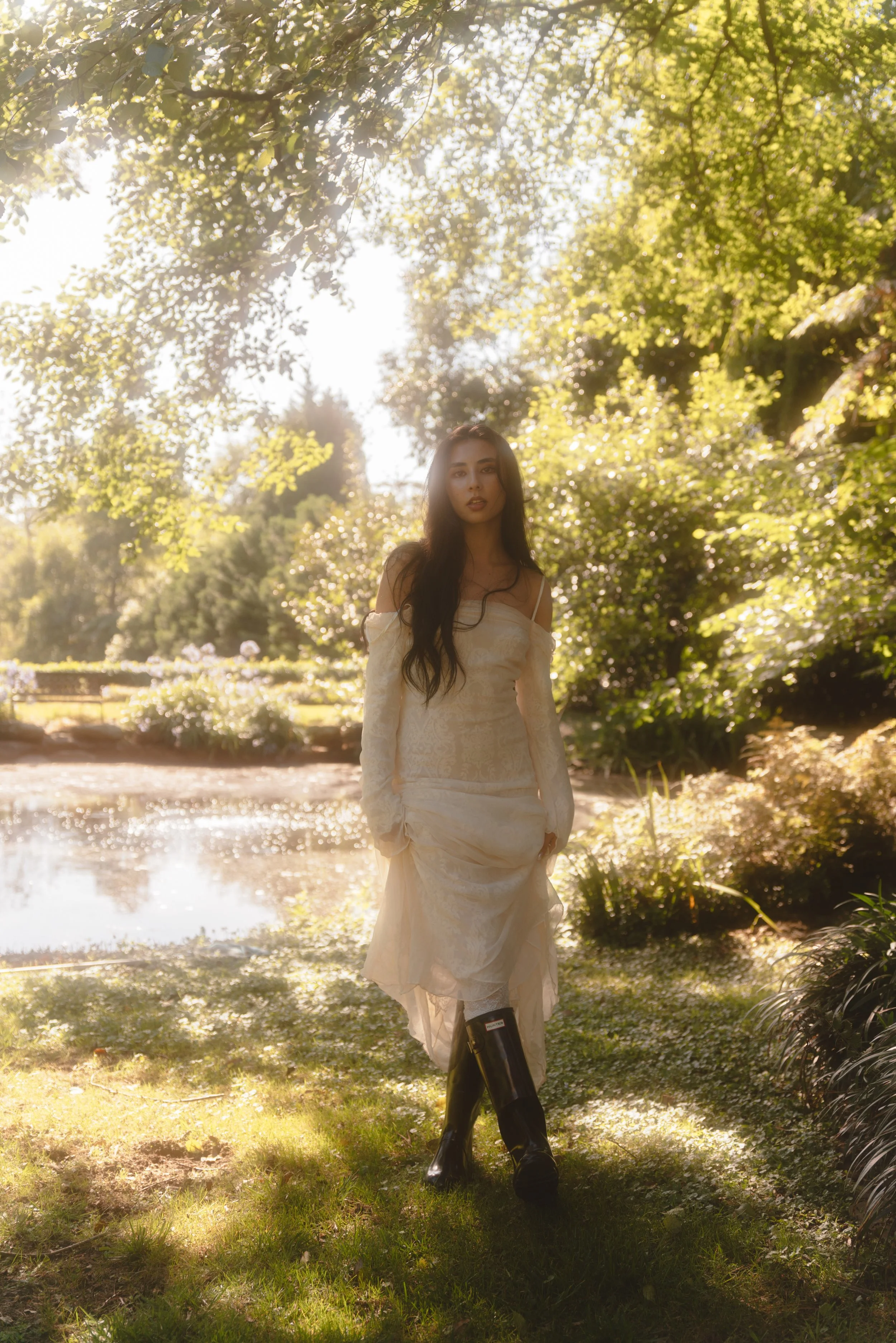 A woman dressed in a white lace dress and black rain boots standing outdoors in a lush, sunlit garden with a pond in the background.