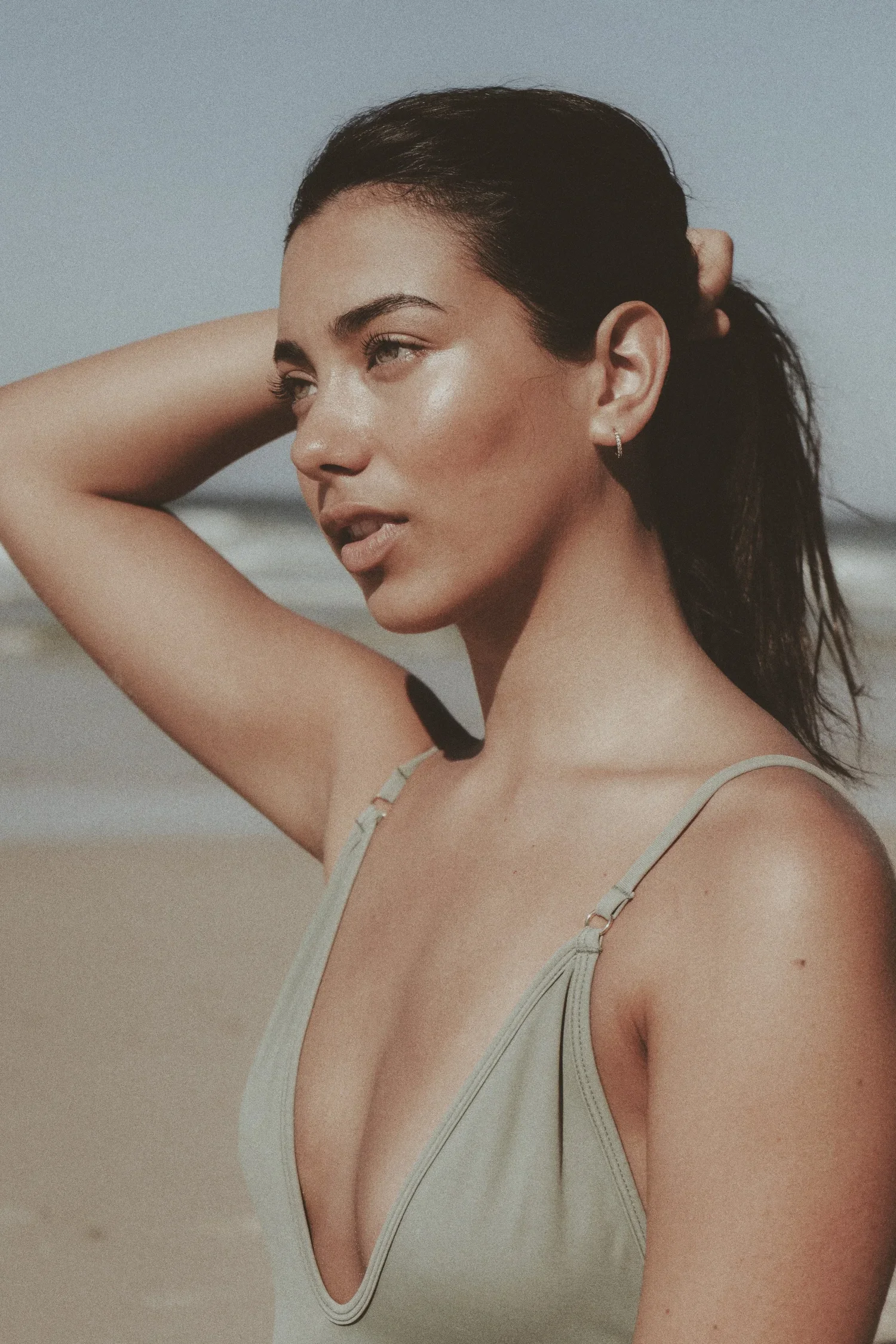 A woman with dark hair in a ponytail, wearing a beige swimsuit, standing on a beach with ocean waves in the background.