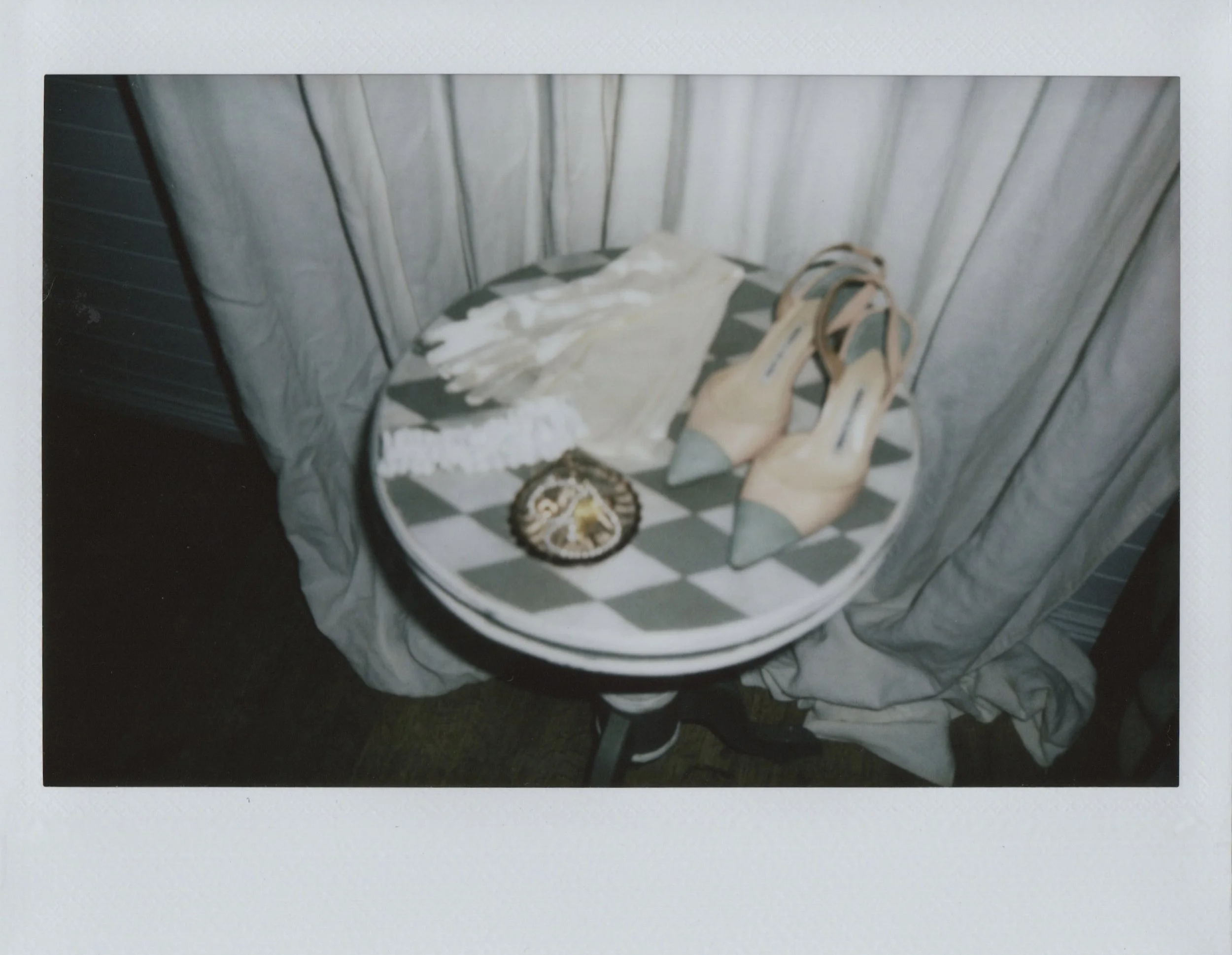 A round table with black and white checkered pattern, set against a curtain. On the table are a white lace garment, a small circular object, and a pair of high-heeled sandals with gray straps and beige heels.