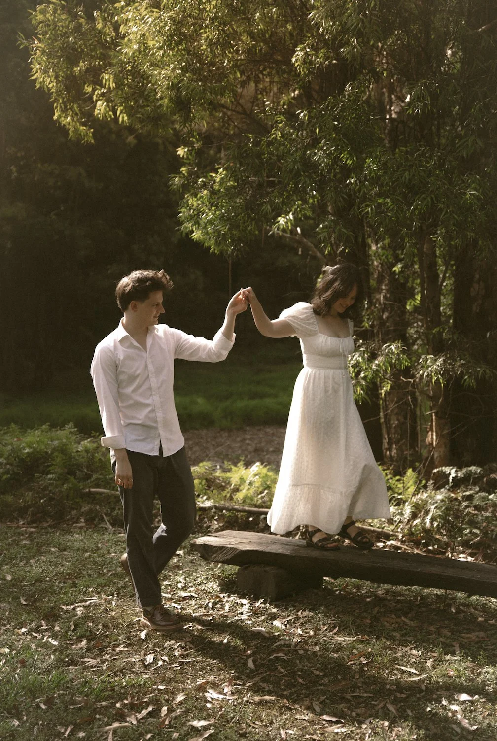 A man and woman dance outdoors at sunset, with the man helping the woman walk across a narrow wooden bridge, surrounded by trees and greenery.