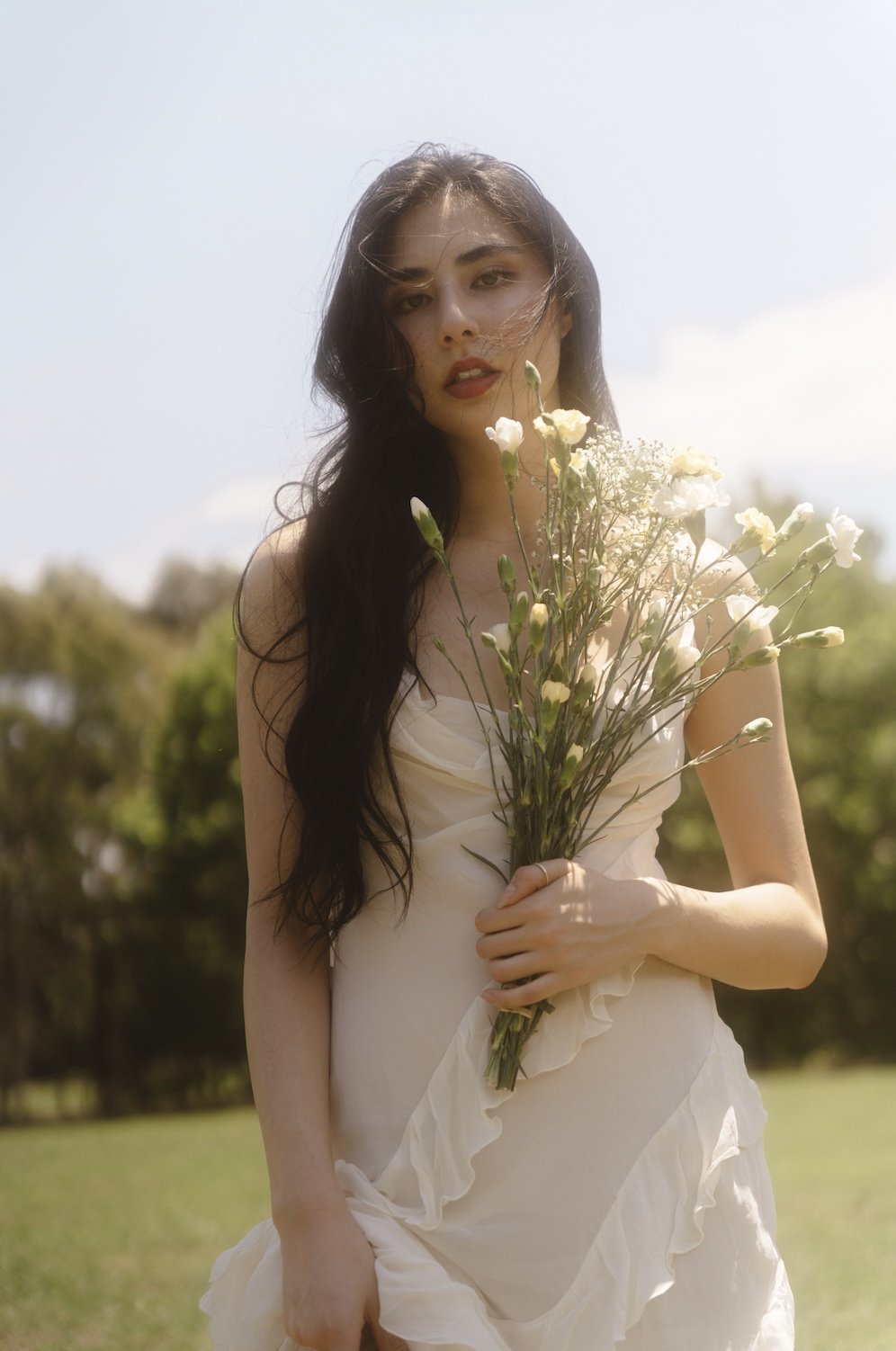 A young woman with long dark hair holding a bouquet of white flowers outdoors on a sunny day.